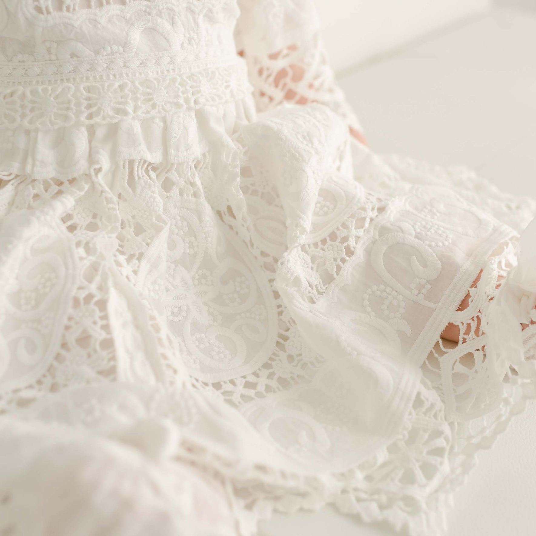 Close-up of a white lace fabric, showcasing detailed floral and paisley patterns. The intricate lace texture, often seen in an Adeline Lace Christening Dress & Bloomers, features various designs with a scalloped edge visible at the bottom. The image highlights the delicate and ornate nature of the fabric.