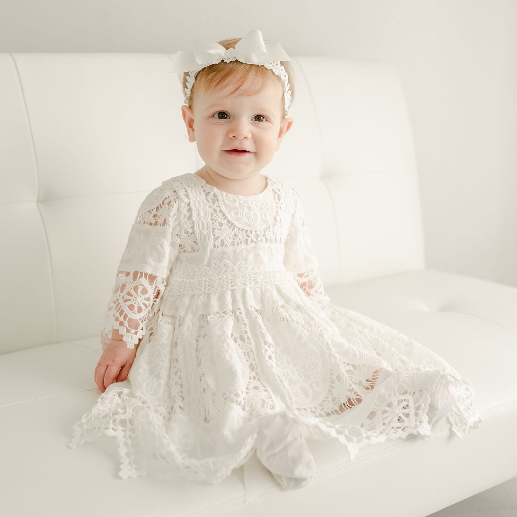 A young child with light hair sits on a white couch, wearing the elegant Adeline Lace Christening Dress & Bloomers, complemented by a matching white headband with a bow. The outfit is finished with a delicate silk ribbon sash. The child looks at the camera against a plain white background.