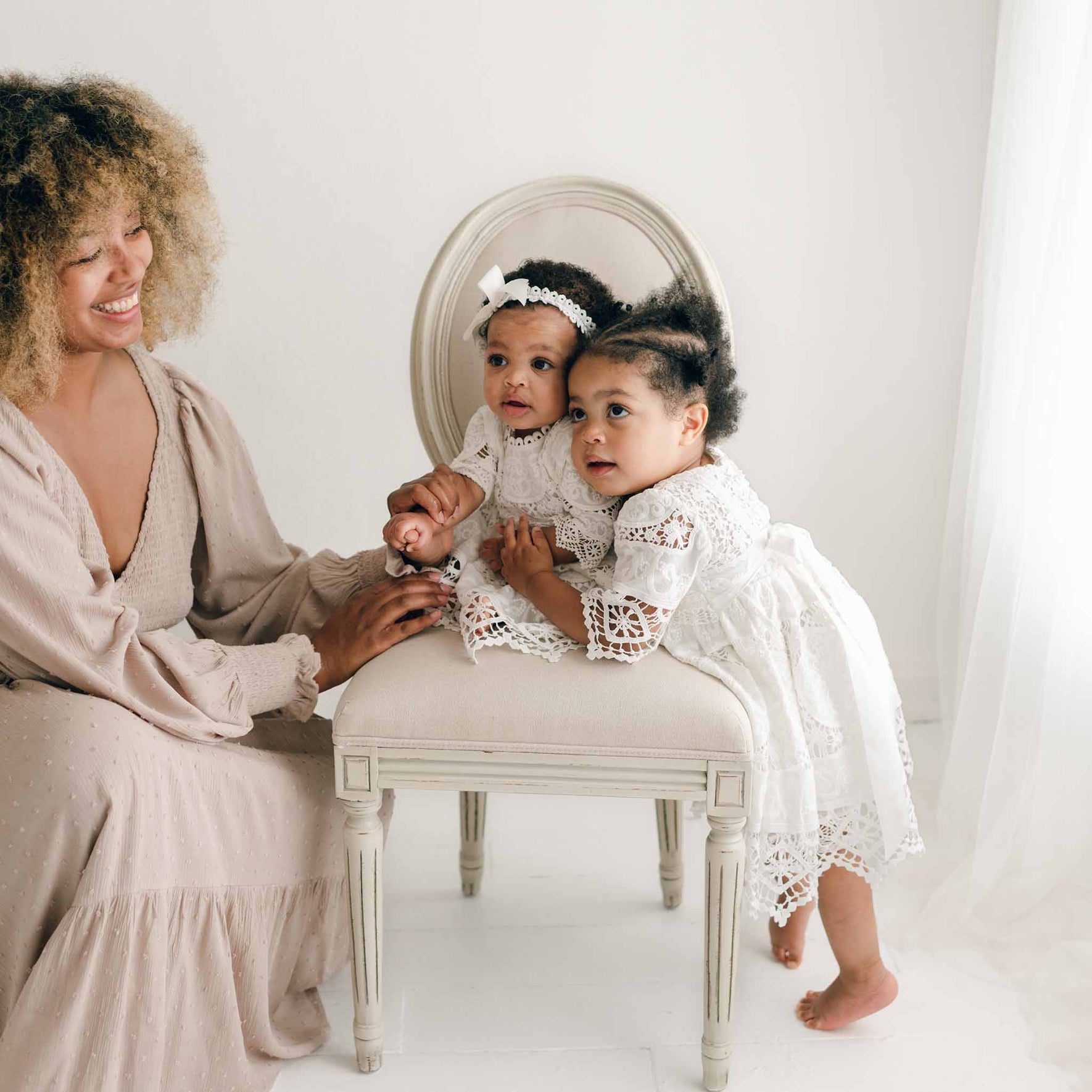 A woman in a long, beige dress sits beside a cushioned chair. Two young girls in matching Adeline Lace Christening Dresses & Bloomers are on the chair, with one standing and the other sitting with a white headband and a silk ribbon sash. The scene is set in a bright room with white walls and curtains.