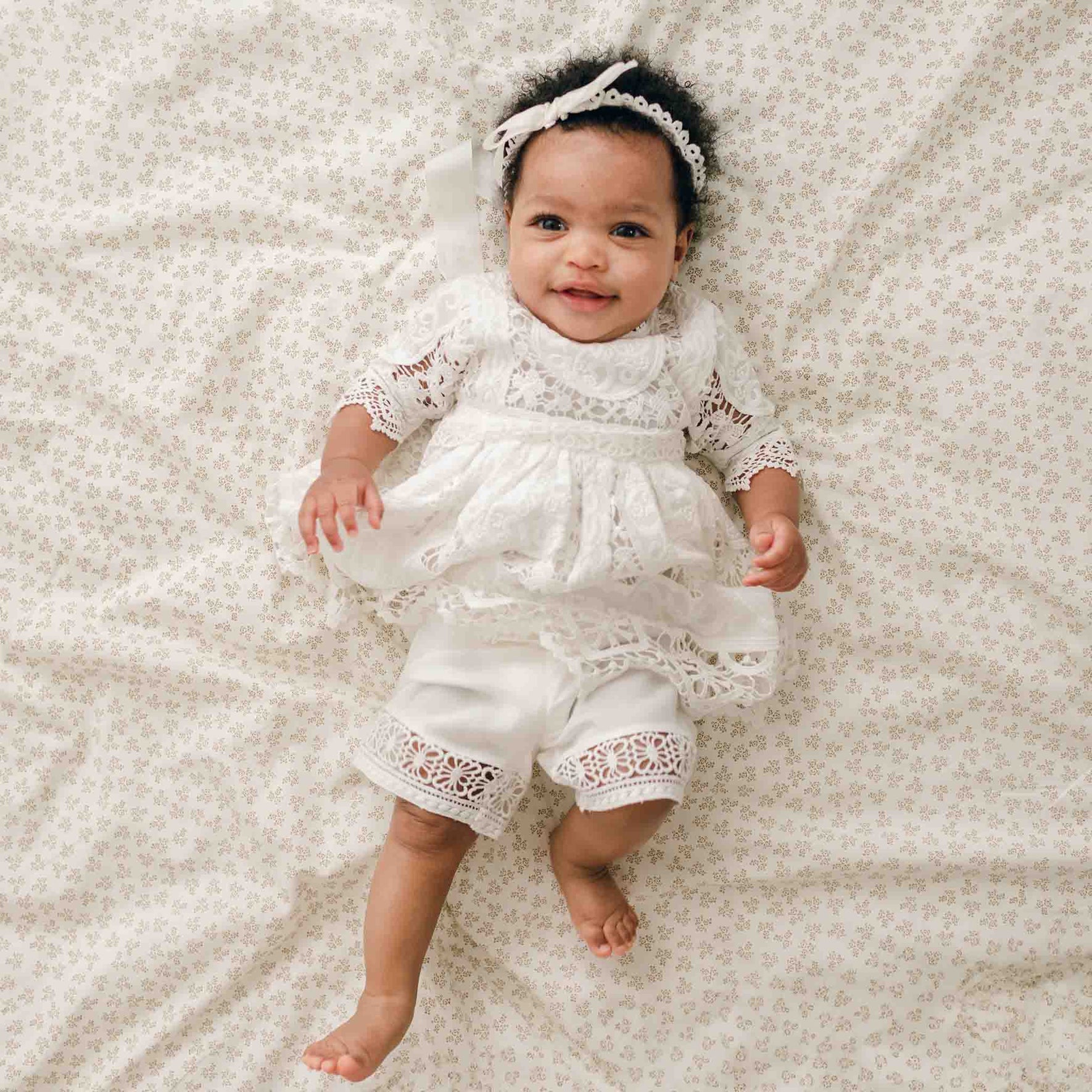 A baby lies on a patterned blanket, wearing the Adeline Lace Christening Dress & Bloomers with a matching headband. The silk ribbon sash adds an elegant touch as the baby looks at the camera and smiles.