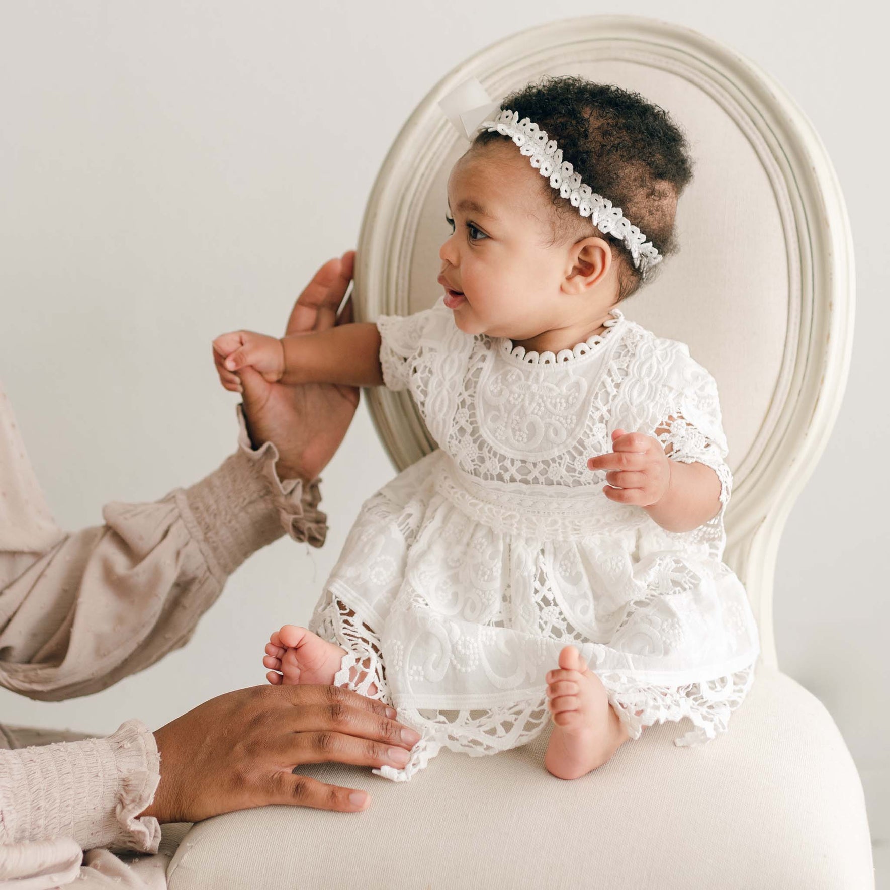A baby wearing the Adeline Lace Christening Dress & Bloomers, featuring a white lace design with a silk ribbon sash and accessorized with a headband adorned with a bow, sits on a beige chair. An adult's hands lightly support the baby, who looks to the side against a plain, light-colored background.