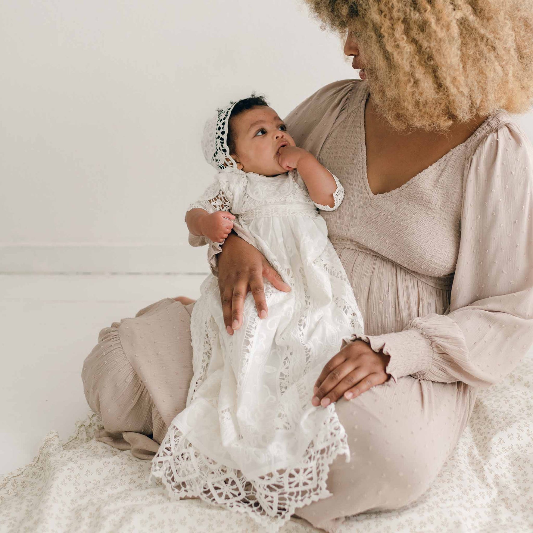 A woman with curly hair sits on the floor, facing a baby dressed in an Adeline Lace Christening Gown & Bonnet, who rests on her lap. The woman is wearing a light pink dress with long sleeves. The baby, adorned in beautiful cotton lace, looks up at the woman while sucking on its thumb.
