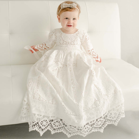 A young child sits on a white sofa, wearing the Adeline Lace Christening Gown & Bonnet. The elegant christening gown and matching bonnet frame the child's gentle smile as they look forward. The neutral background enhances the beauty of the heirloom set and captures the child's serene expression perfectly.