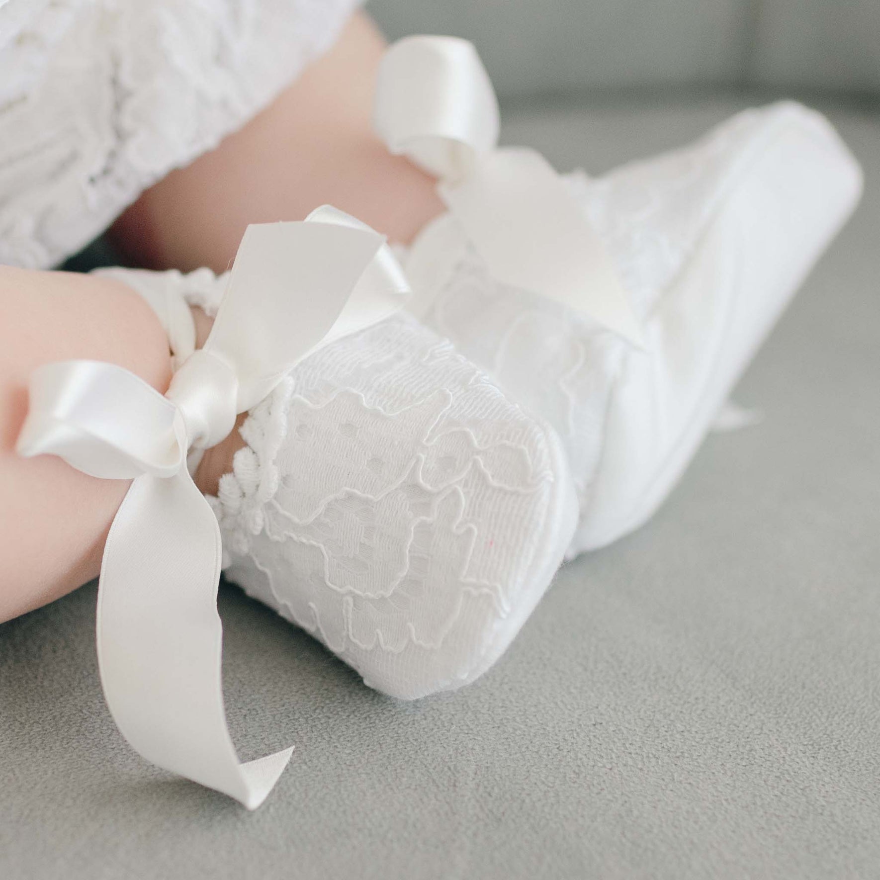 A close-up of a baby wearing delicate white lace shoes with large satin bows, part of the Aria Accessory Bundle - 15%, ideal for baptisms. The floral embroidered pattern beautifully complements the soft, light-colored surface beneath the baby.