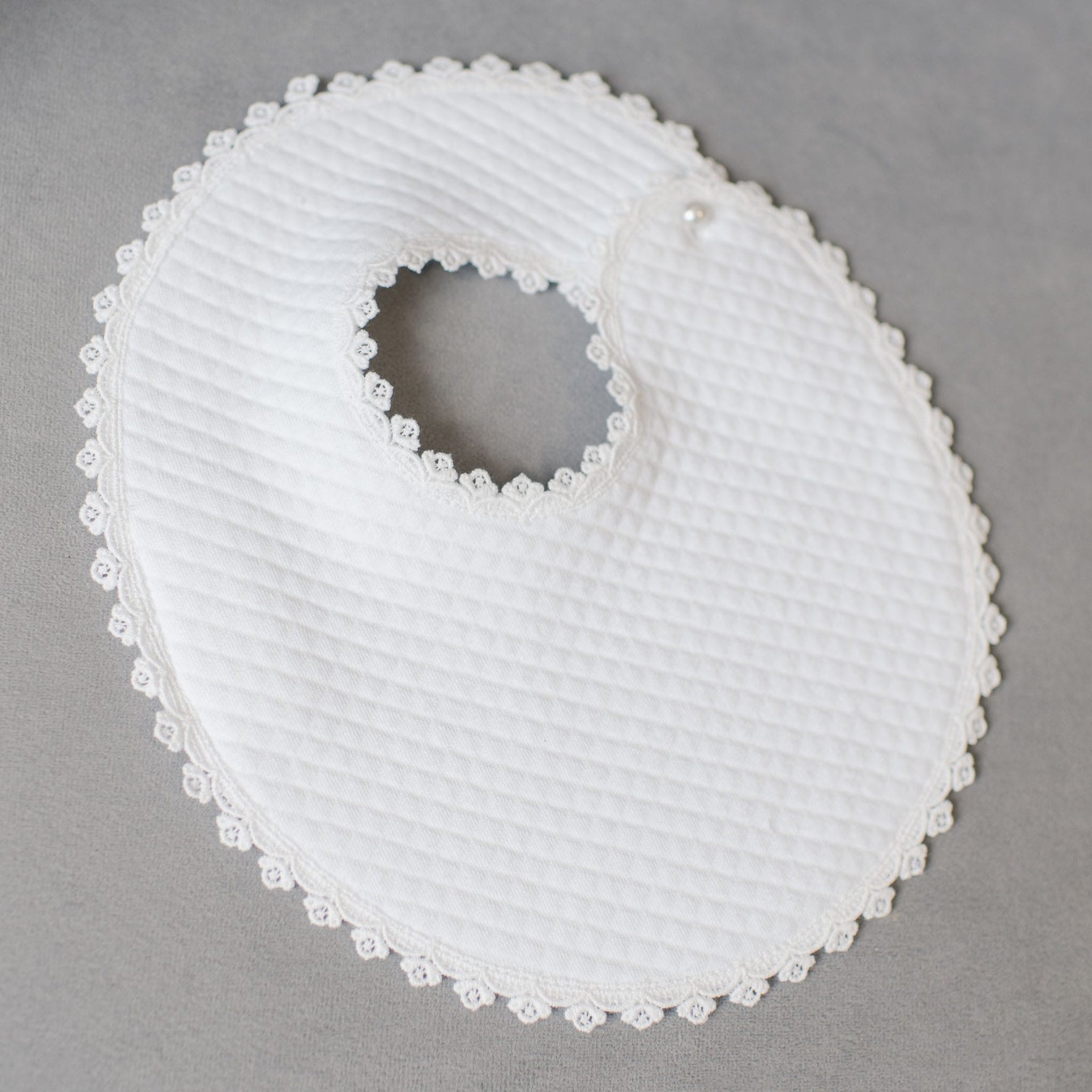 The Aria Bib is displayed on a gray surface, showcasing its round shape and white textured fabric complemented by a trim of Venice lace. The bib exudes the elegance of a christening gown, with a circular neck opening and a small decorative button near the closure, capturing the essence of designer baby attire.