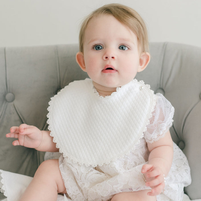 A baby with blue eyes and light brown hair sits on a gray tufted chair, wearing a christening gown adorned with Venice lace. The outfit, complemented by an Aria Bib and a white lace dress, blends in beautifully with the softly lit neutral background.