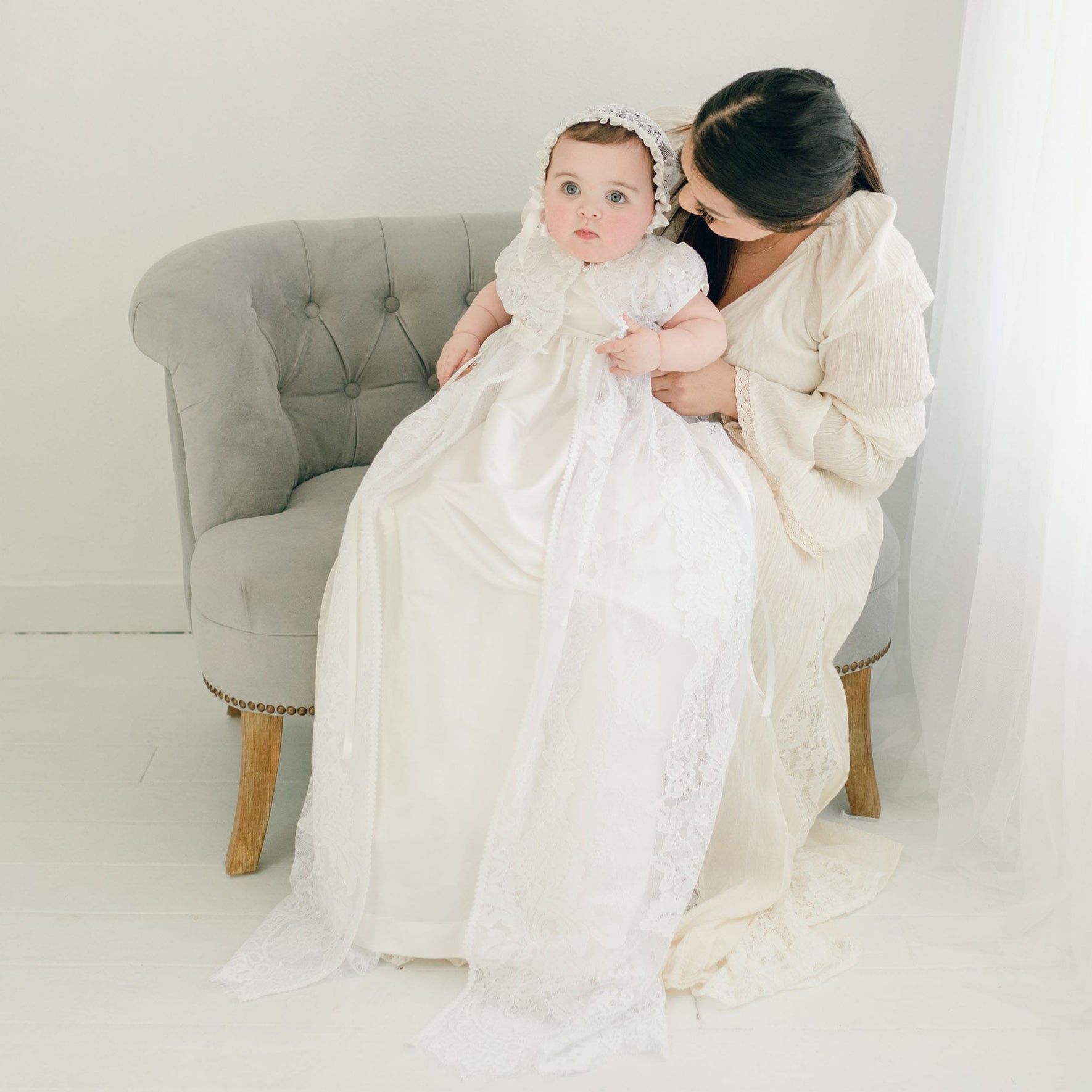 A woman sits on a grey tufted chair, gently holding a baby dressed in the Aria Christening Gown & Bonnet. She wears a cream-colored dress as they sit near a bright window, bathed in a soft glow that highlights the cherished moment.