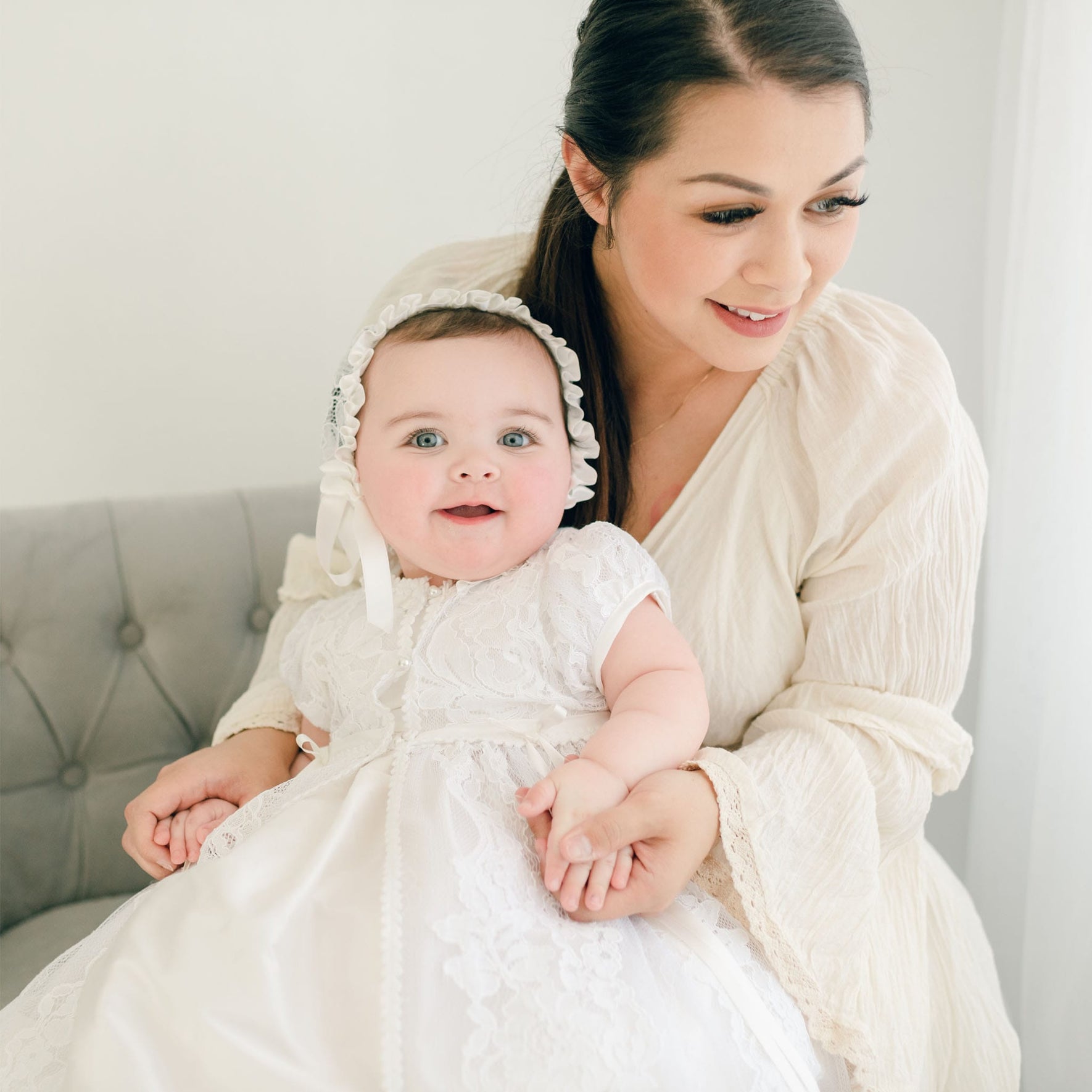 A woman in a light-colored dress holds a smiling baby dressed in the exquisite Aria Christening Gown & Bonnet, as they sit on a gray cushioned seat against a softly lit background.