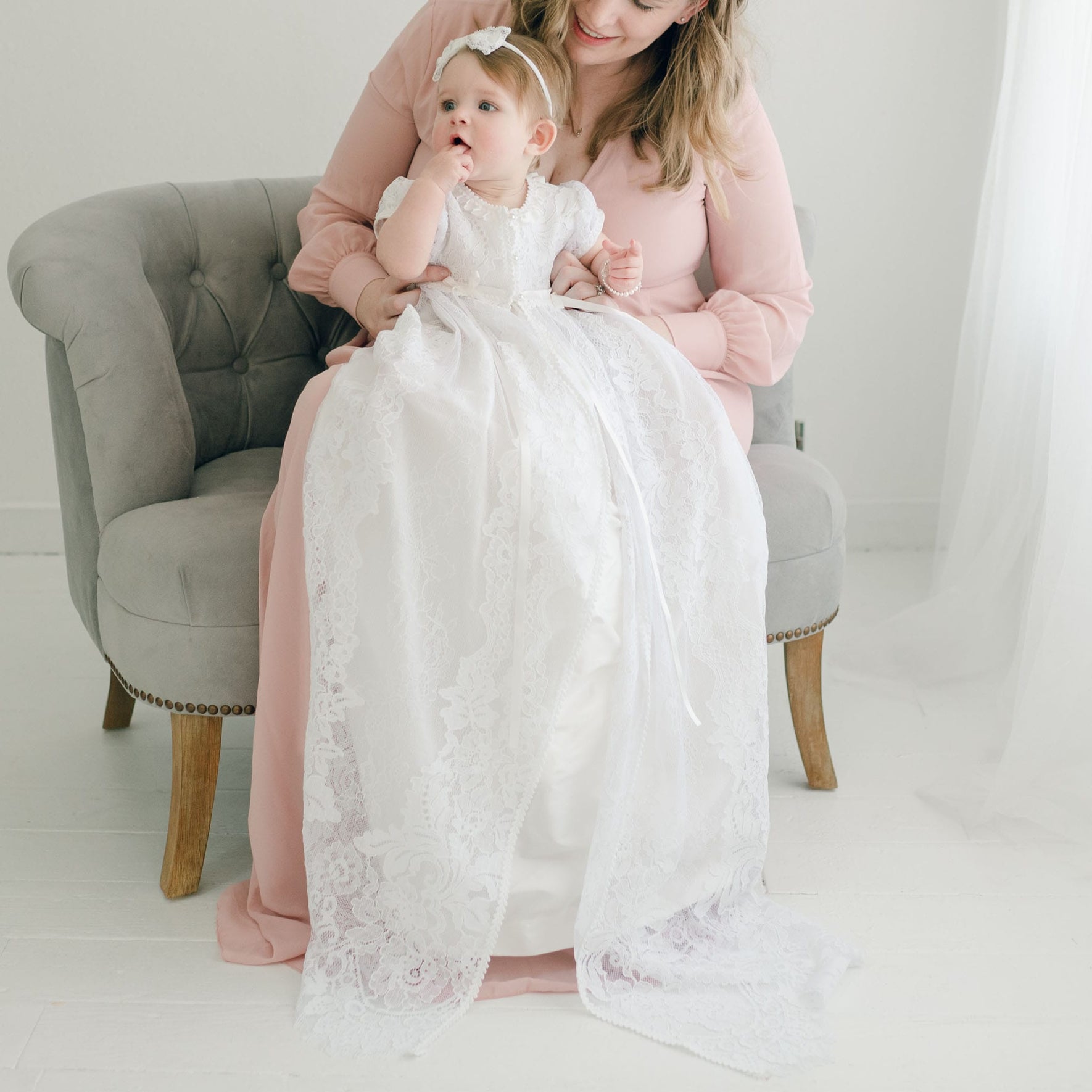 A woman in a pink dress sits on a gray armchair, cradling a baby dressed in the Aria Christening Gown & Bonnet, adorned with lace details. The baby, wearing a delicate headband, gazes to the side while the woman's face is partially visible and smiling. The softly lit setting exudes elegance.