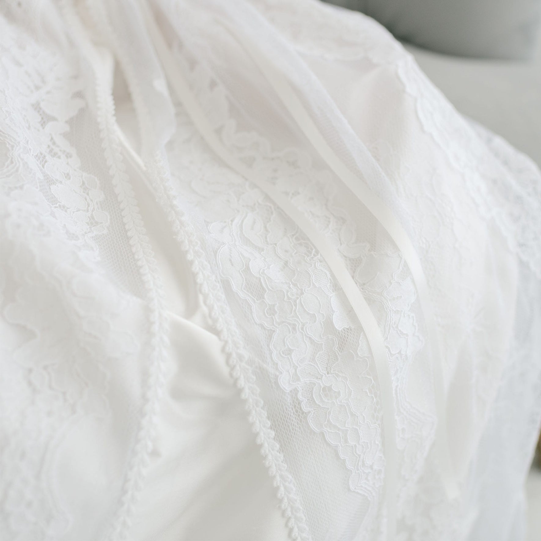 Close-up of a white lace fabric with intricate floral patterns and delicate ribbon details, evocative of an heirloom piece. The fabric seems to be part of the Aria Christening Gown & Bonnet, softly draped in elegant folds.