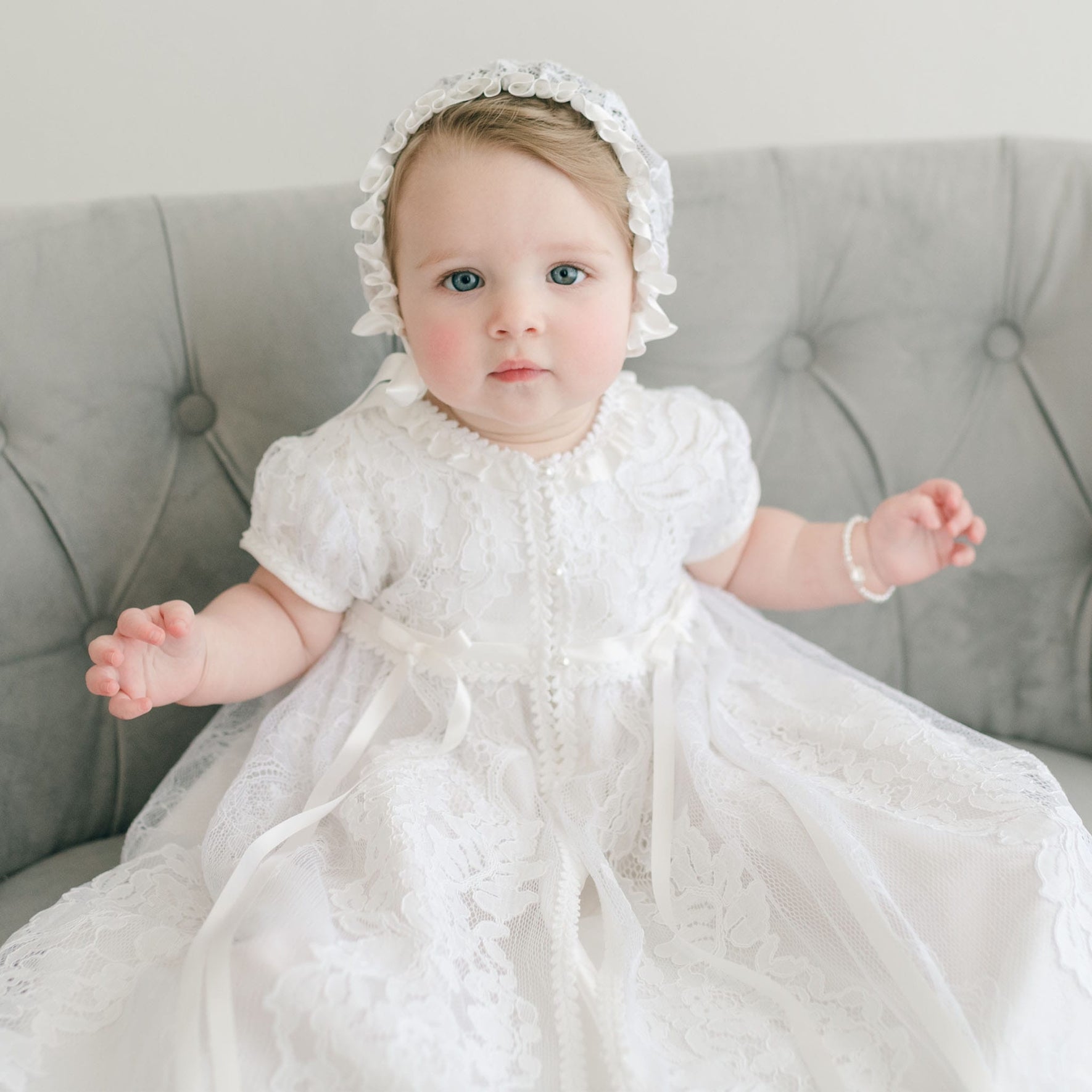 A baby dressed in the exquisite Aria Christening Gown & Bonnet, featuring a white lace dress, sits on a gray tufted couch. The child, with light-colored hair and eyes, also wears a small bracelet on the left wrist. A plain light-colored wall serves as the backdrop.
