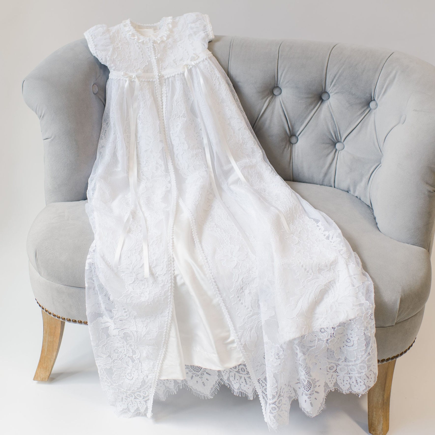 A long, heirloom piece of the Aria Christening Gown & Bonnet is draped over a light gray, tufted loveseat with wooden legs. The gown showcases intricate lace patterns and short sleeves, spread out to display its full length and detail, against a neutral background.