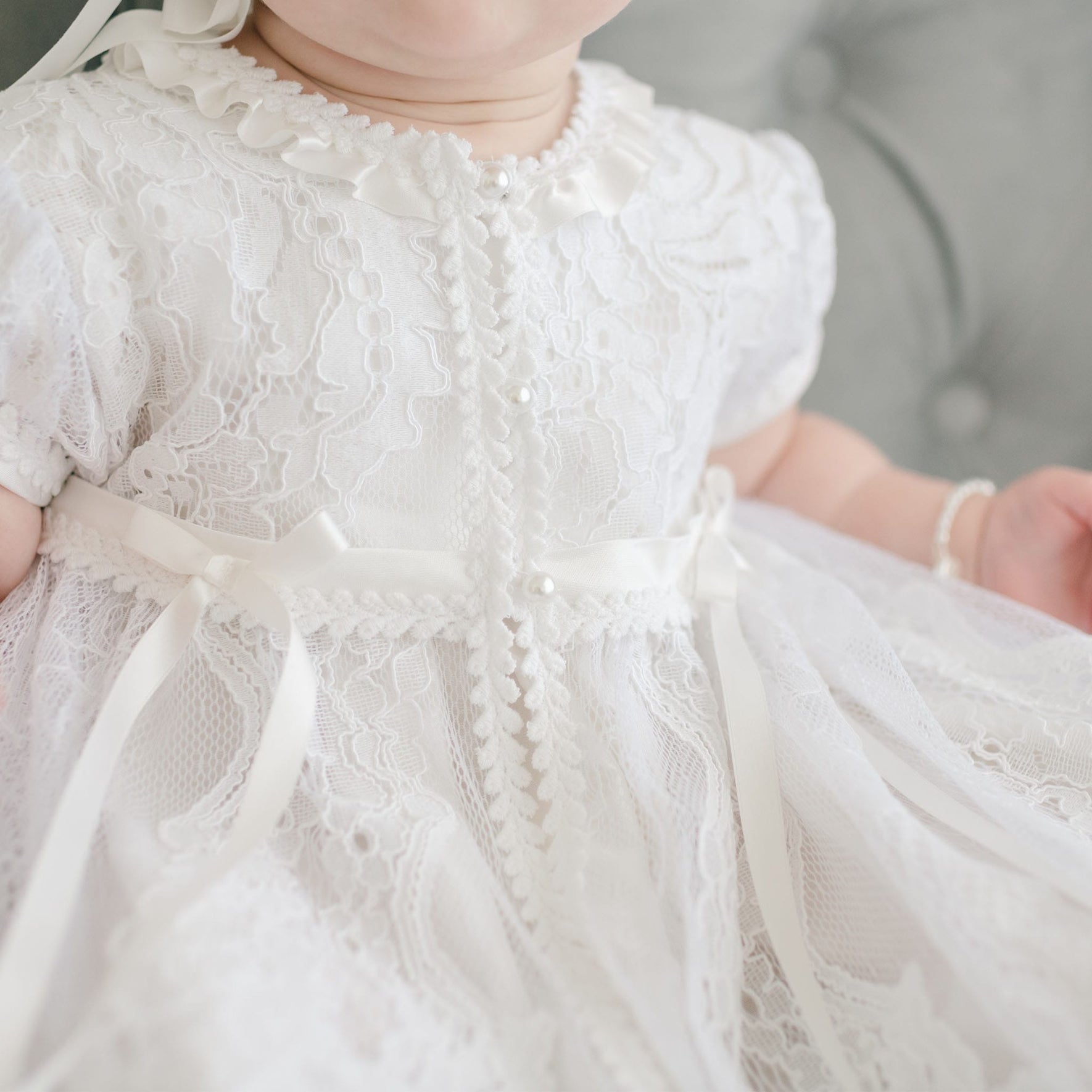 A baby wearing the Aria Christening Gown & Bonnet, adorned with intricate pearl buttons and ribbon accents, sits on a tufted cushion. This heirloom piece, showcasing a ruffled collar and short sleeves, captures a timeless elegance, though the baby's face is partially out of the frame.