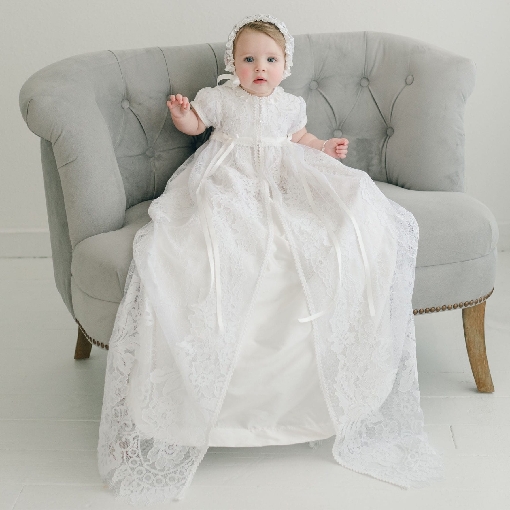 A baby dressed in the Aria Christening Gown & Bonnet sits on a gray upholstered chair. This ornate gown, an heirloom piece, showcases intricate lace patterns. The room is adorned with white walls and a light-colored floor.