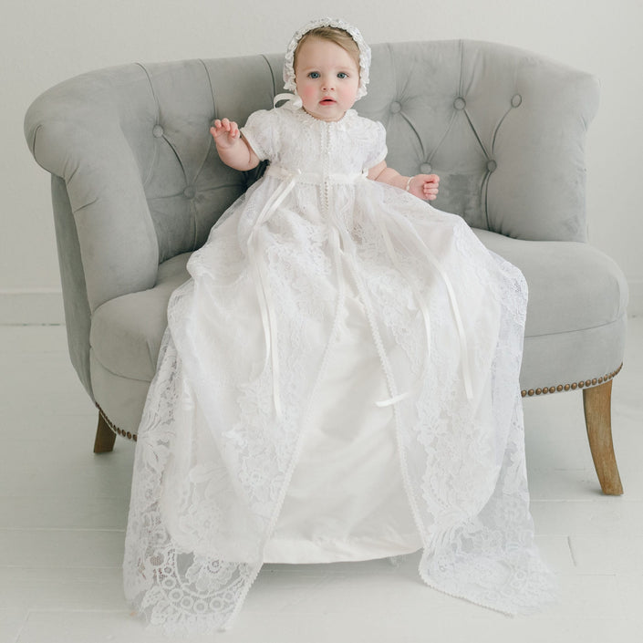 A baby dressed in the Aria Christening Gown & Bonnet sits on a gray upholstered chair. This ornate gown, an heirloom piece, showcases intricate lace patterns. The room is adorned with white walls and a light-colored floor.
