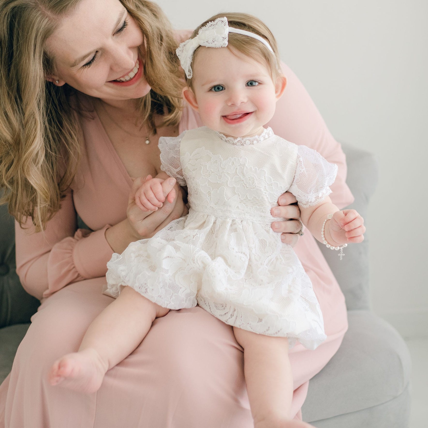 A woman in a light pink dress sits on a gray couch holding a delighted baby dressed in a white lace outfit adorned with the Aria Lace Bow Headband. The baby, wearing a bracelet and holding the woman's fingers, looks content and happy.