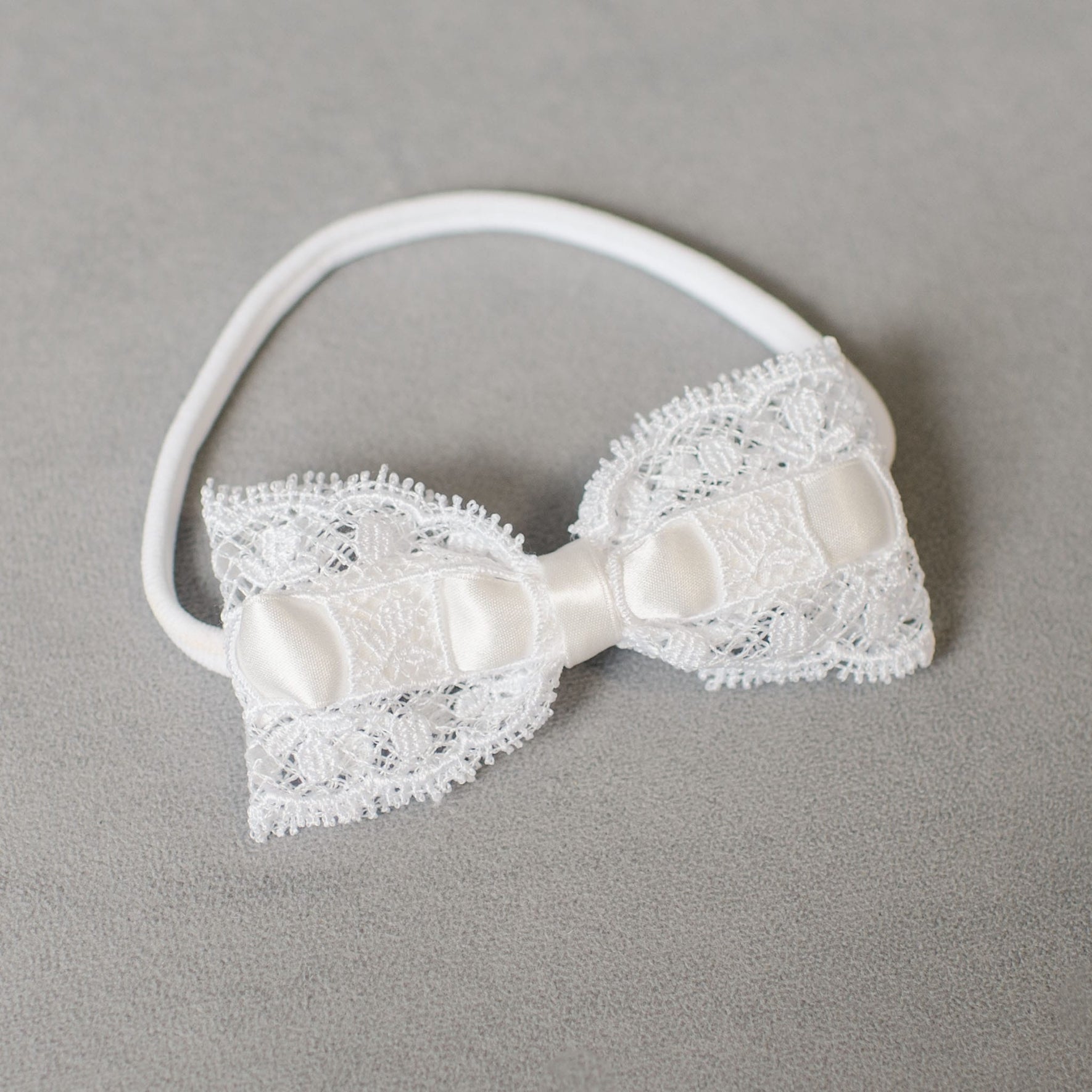 The Aria Lace Bow Headband is showcased on a gray surface, highlighting intricate lace patterns and a hand-stretched nylon base with a satin ribbon accent at the center.