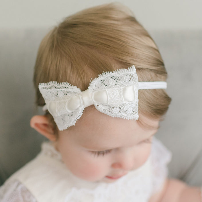 A baby adorning the Aria Lace Bow Headband gazes downward. With light brown hair, the little one is dressed in a charming white lace outfit, exuding elegance and grace.