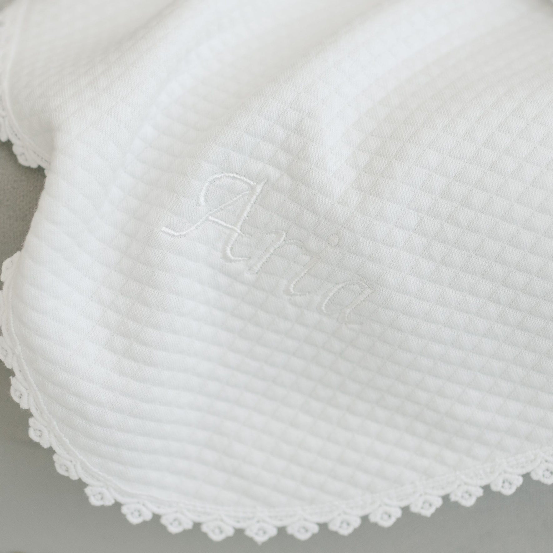 The Aria Personalized Blanket is a white textured fabric with "Aria" embroidered in matching white thread. Featuring a diamond pattern and delicate lace trim along the edge, it's perfect as a christening blanket and makes an ideal personalized gift.