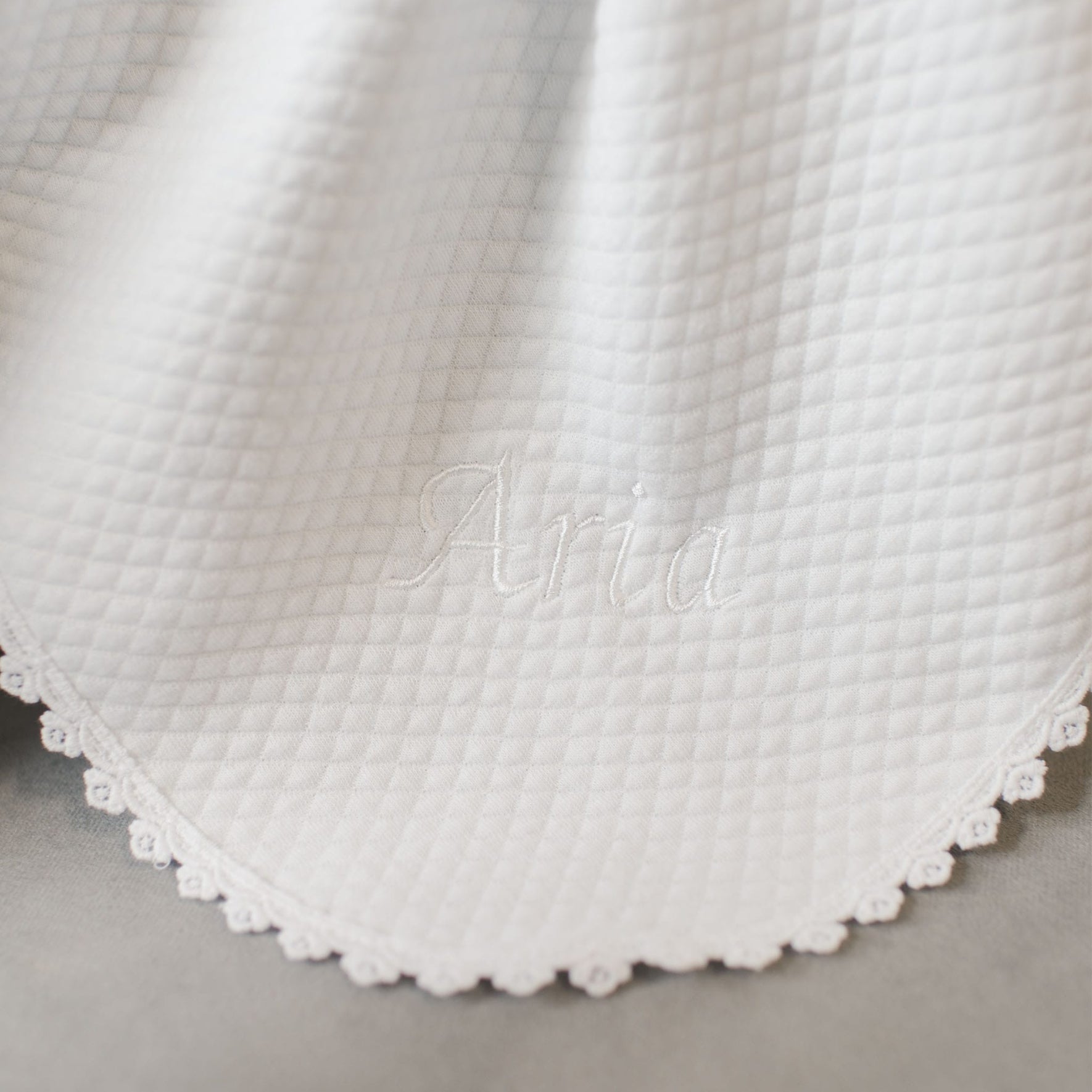 A detailed view of the Aria Personalized Blanket reveals its white, quilted texture and scalloped edges. The name "Aria" is elegantly embroidered in the center with white thread, making it an ideal personalized gift for a baptism outfit.