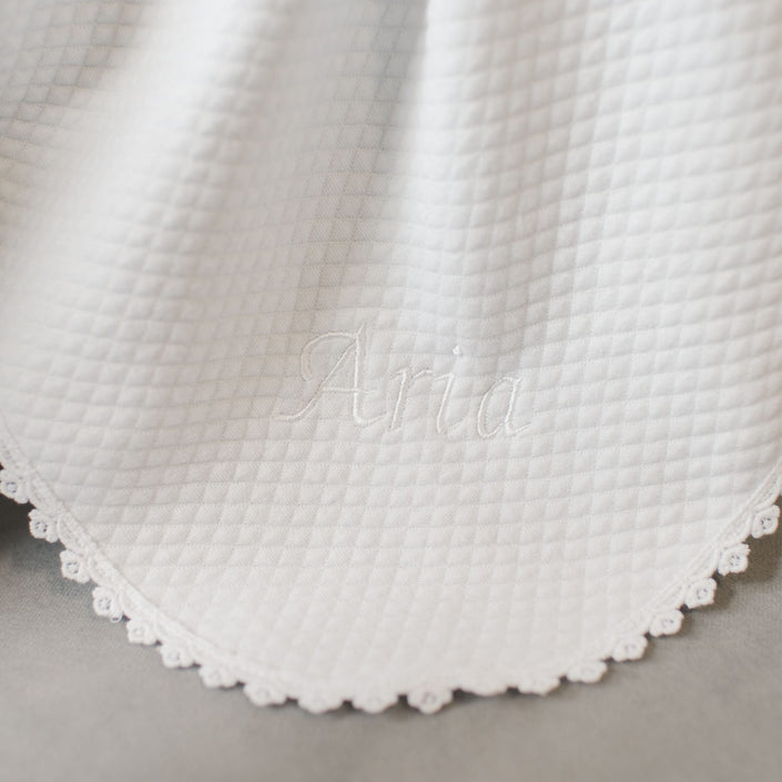 A detailed view of the Aria Personalized Blanket reveals its white, quilted texture and scalloped edges. The name "Aria" is elegantly embroidered in the center with white thread, making it an ideal personalized gift for a baptism outfit.