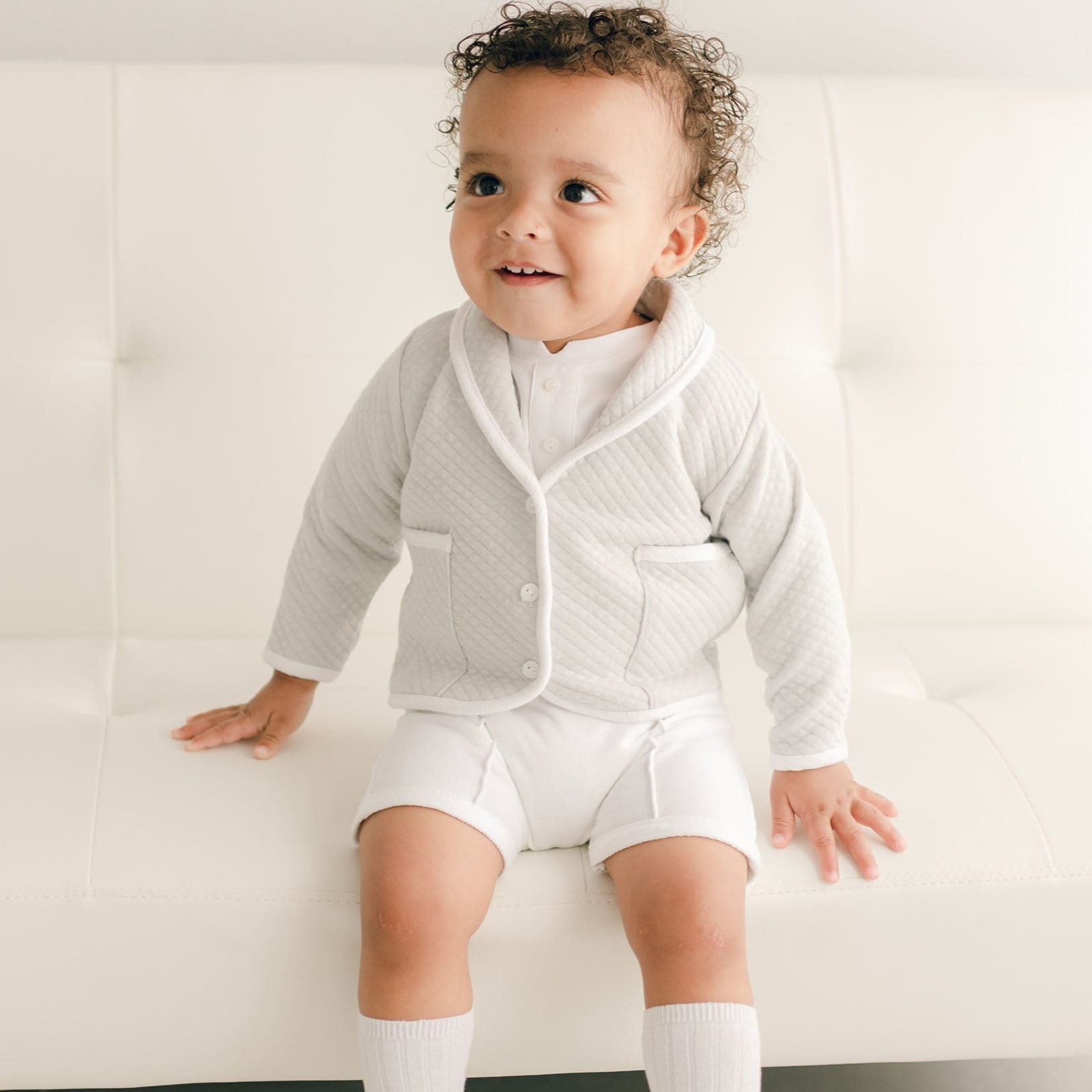 Baby boy sitting on a white couch and wearing the grey Asher 3-Piece Suit, including a folded collar jacket, white pants and a onesie with buttons.