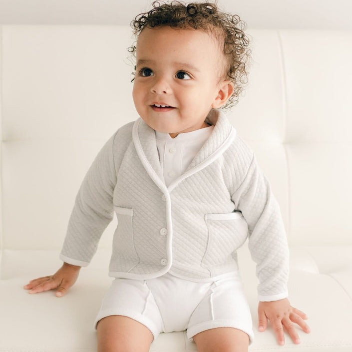 Baby boy sitting on a white couch and wearing the grey Asher 3-Piece Suit, including a folded collar jacket, white pants and a onesie with buttons.