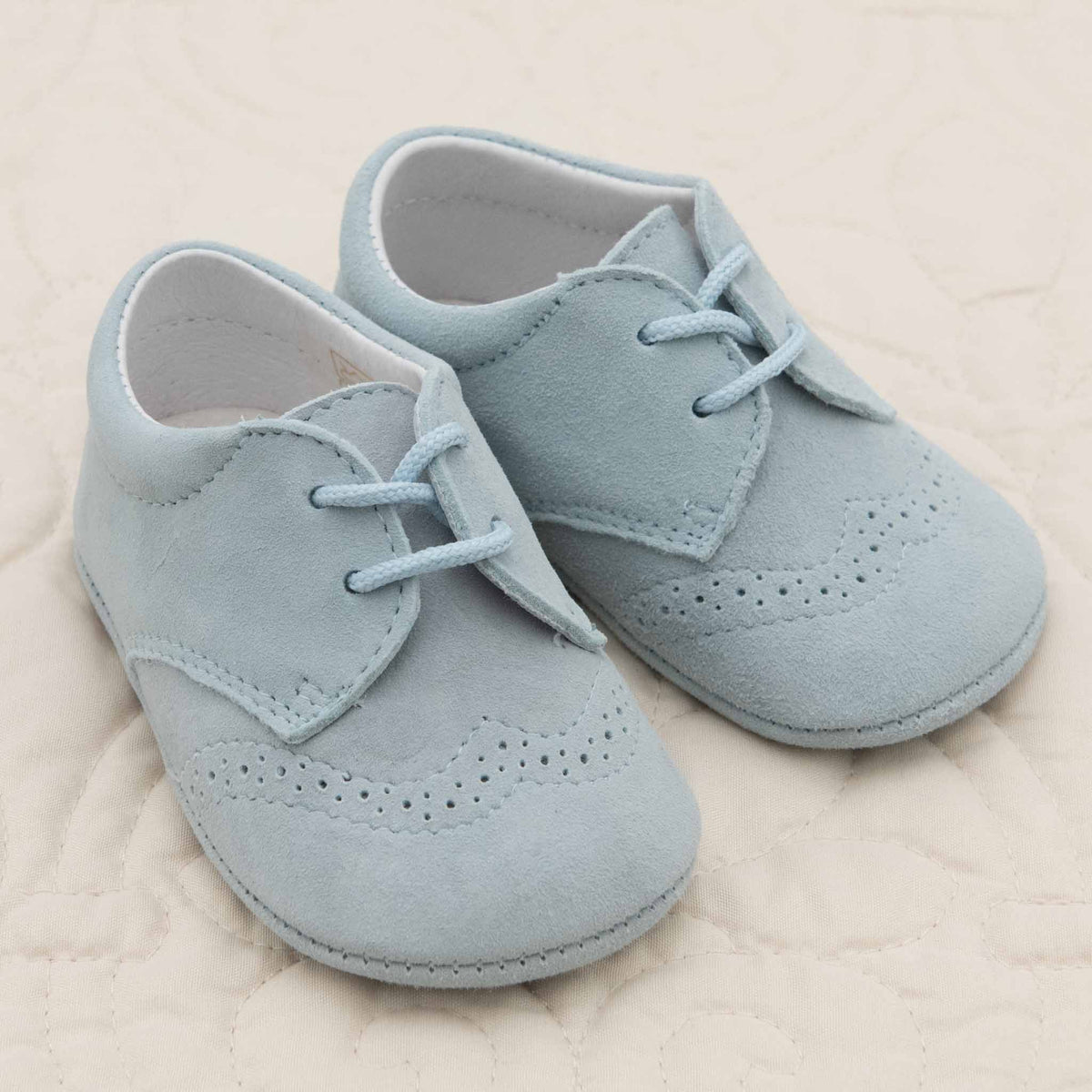 A pair of Asher Suede Shoes in light blue from the Baby Beau & Belle collections, featuring white interiors, lace-up fronts, and decorative stitching on the toes, placed on a beige quilted surface.