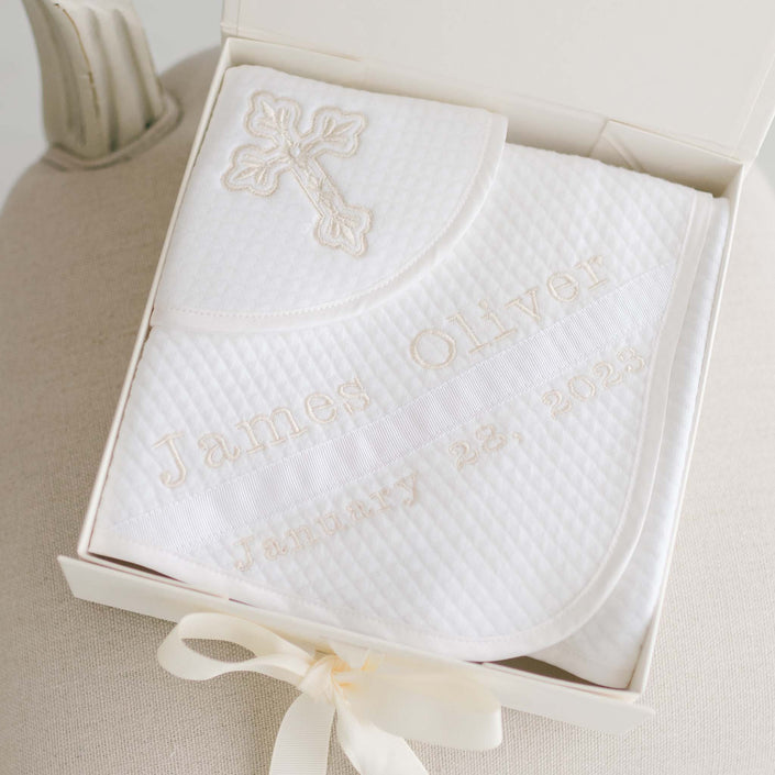 Introducing the Baby Boy Blessing Gift Set: a personalized white quilted cotton blanket elegantly embroidered with a cross and "James Oliver January 28, 2023," presented neatly folded in an open box accented with a ribbon.