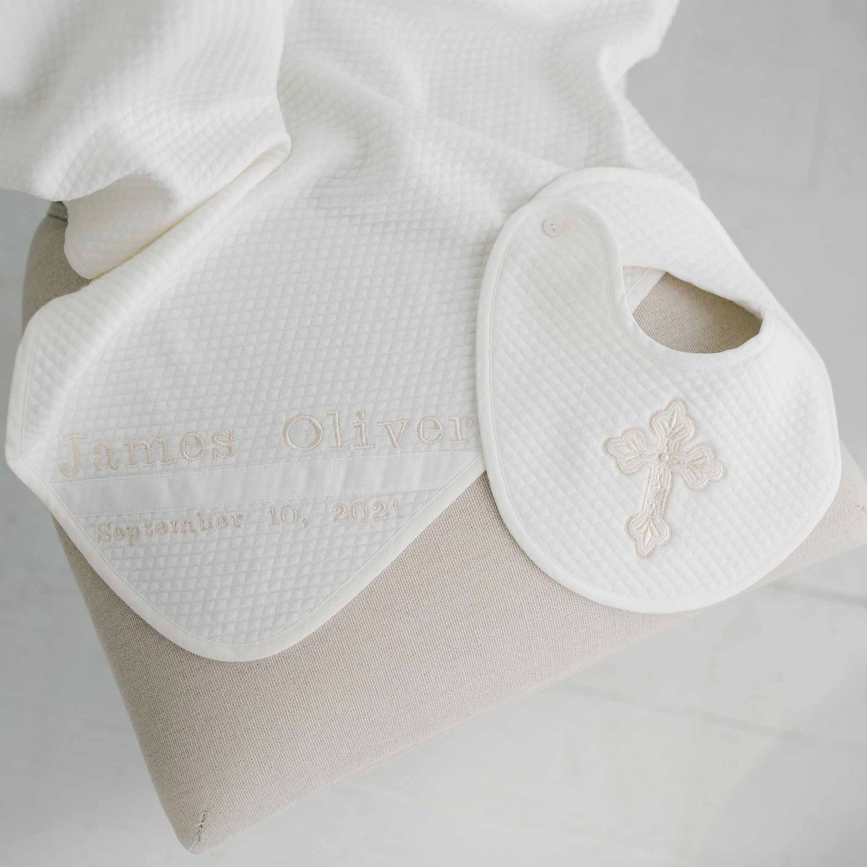 A cozy beige chair is adorned with the Baby Boy Blessing Gift Set, which includes a white bib embroidered with a cross and a quilted cotton blanket featuring the name "James Oliver" and the date "September 10, 2023.