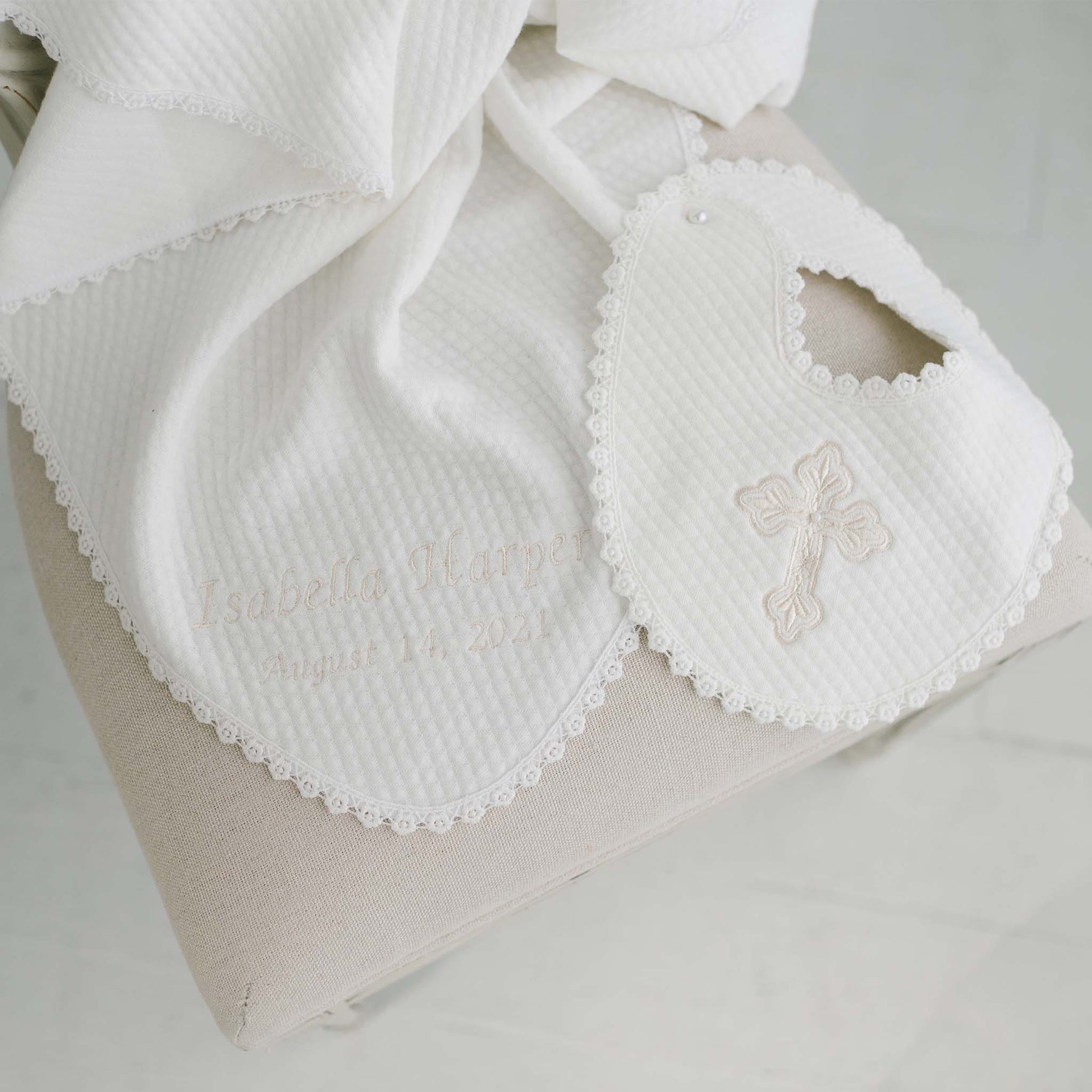 The Baby Girl Blessing Gift Set, perfect as a baptism keepsake, includes a white bib and blanket set featuring a textured pattern and lace trim. The bib is decorated with an embroidered cross, while the personalized gift set contains a baby blanket elegantly stitched with "Isabella Harper" and "August 14, 2021".