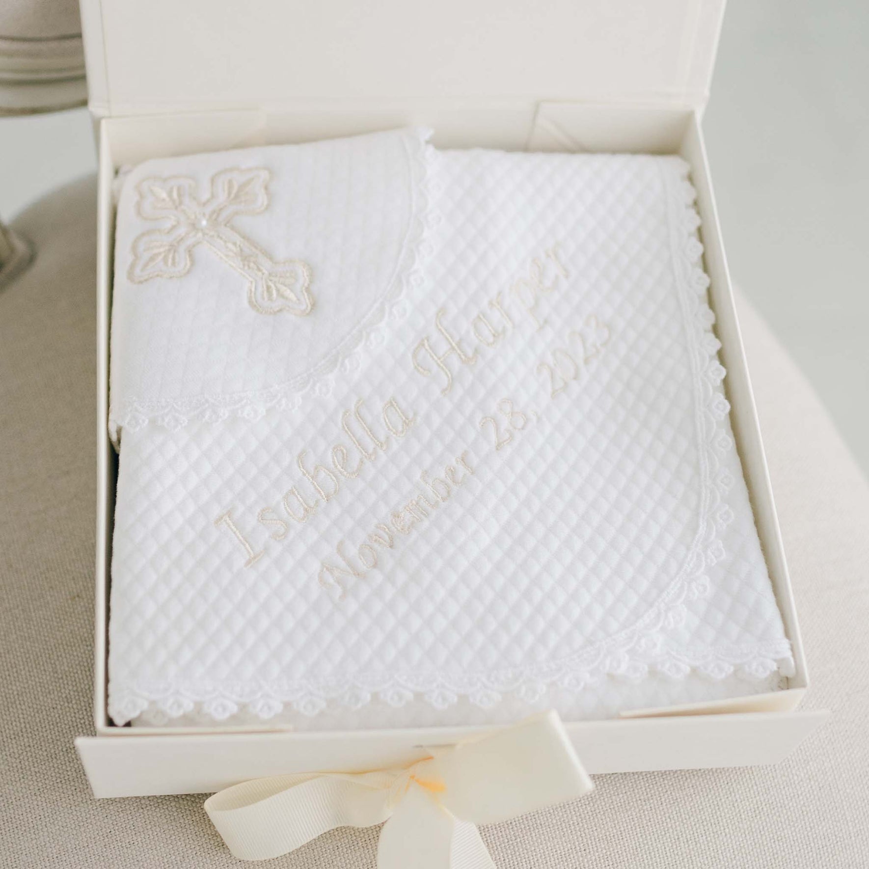 The Baby Girl Blessing Gift Set includes a white, quilted blanket adorned with a cross and personalized with the name "Isabella Harper" and the date "November 28, 2023." Neatly folded in a box tied with a ribbon, it makes an ideal baptism keepsake for your little one.