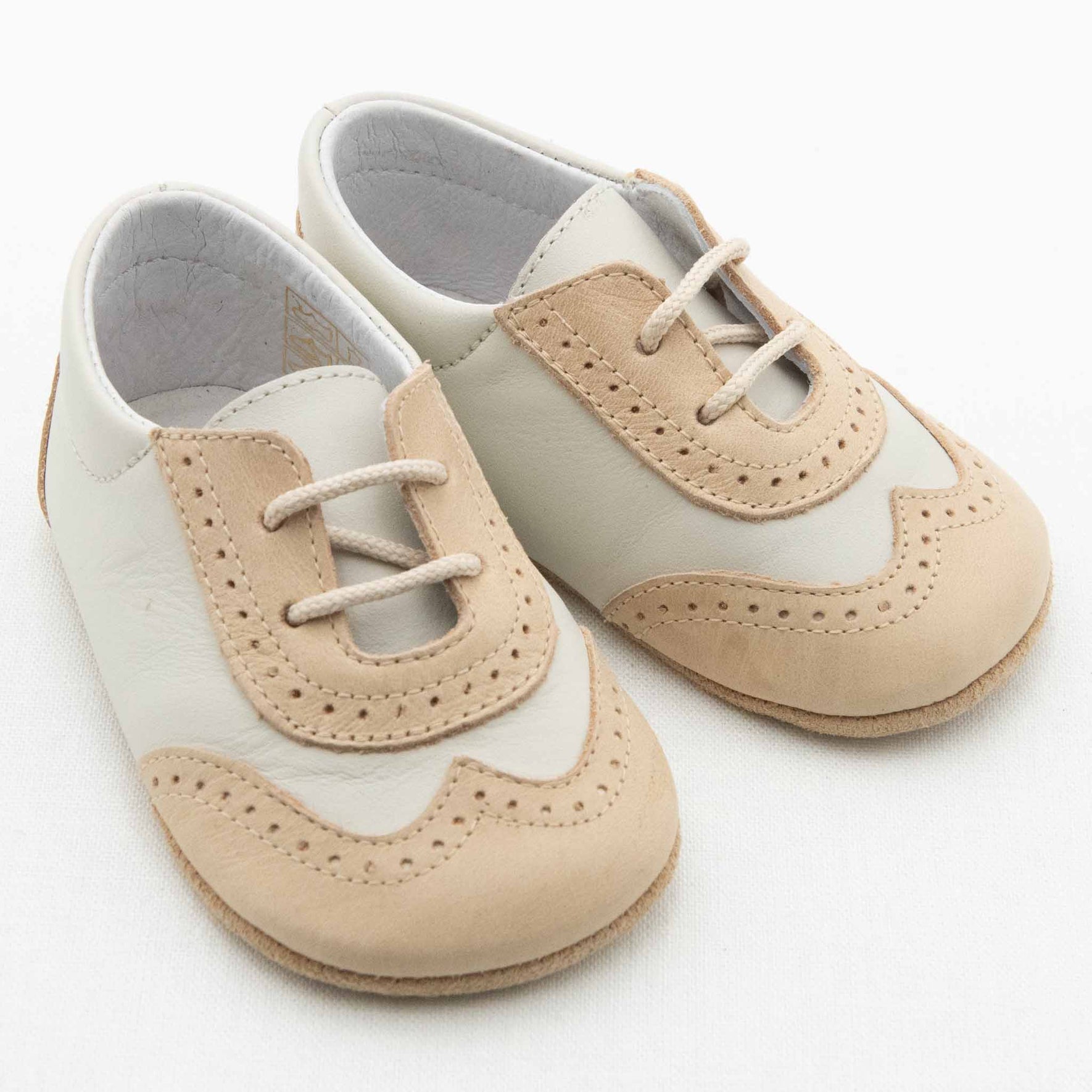 The Boys Beige & Ivory Wingtip Shoes by Baby Beau & Belle are handmade leather baby shoes that feature a creamy white base color with beige accents, laces, and decorative stitching. They come with a soft, flexible sole designed for infants and are presented against a plain white background.
