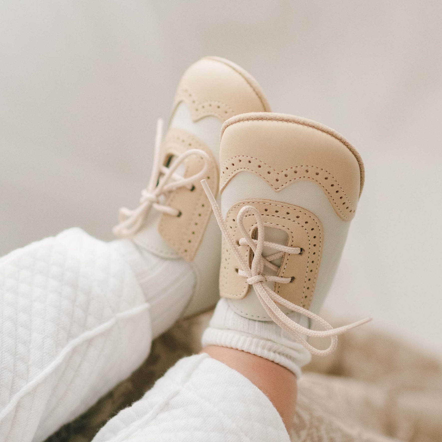 Baby wearing a pair of the Beige and Ivory Wingtip Shoes made with a tan and ivory matte leather with tan suede soles and detailed edging
