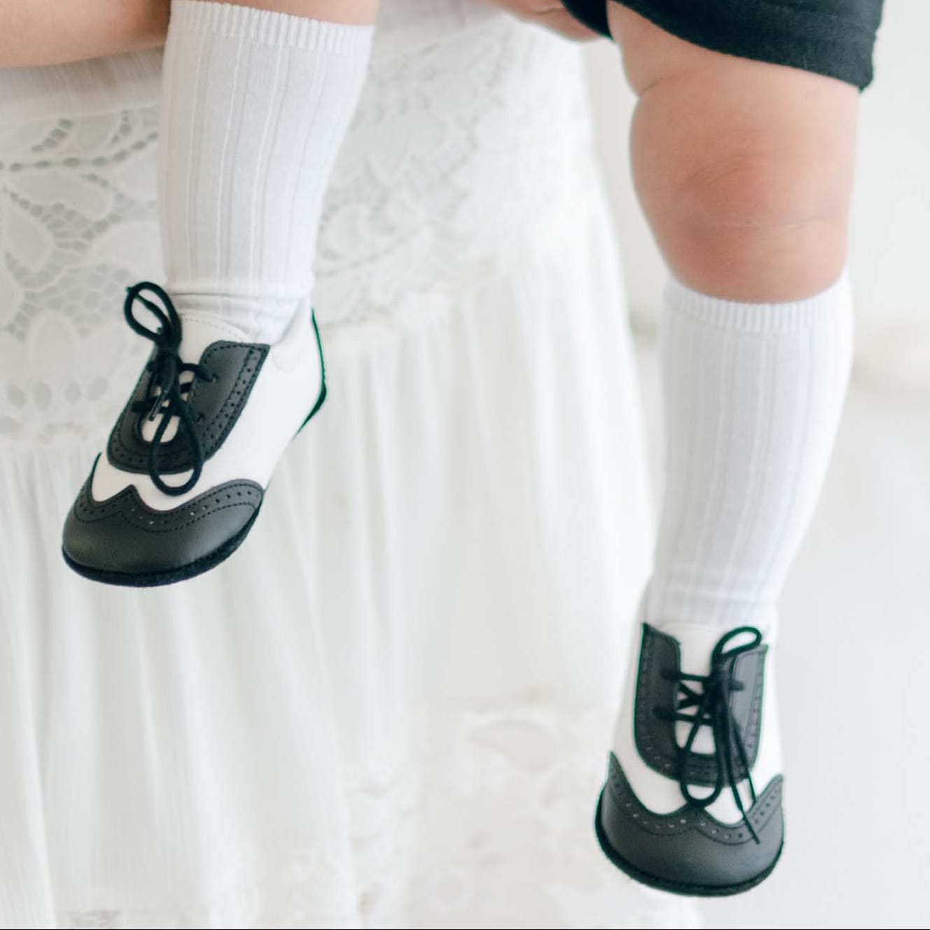 The lower half of a baby boy being held by his mother. He is wearing the James-3 Piece Suit and matching Black and White Leather Wingtip Shoes (with ribbed knee socks).