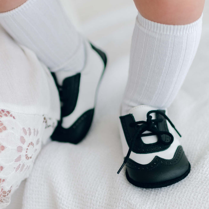 The legs of a baby boy standing on white textured fabric. He is wearing Black and White Leather Wingtip Shoes with matching White Ribbed Knee Socks.
