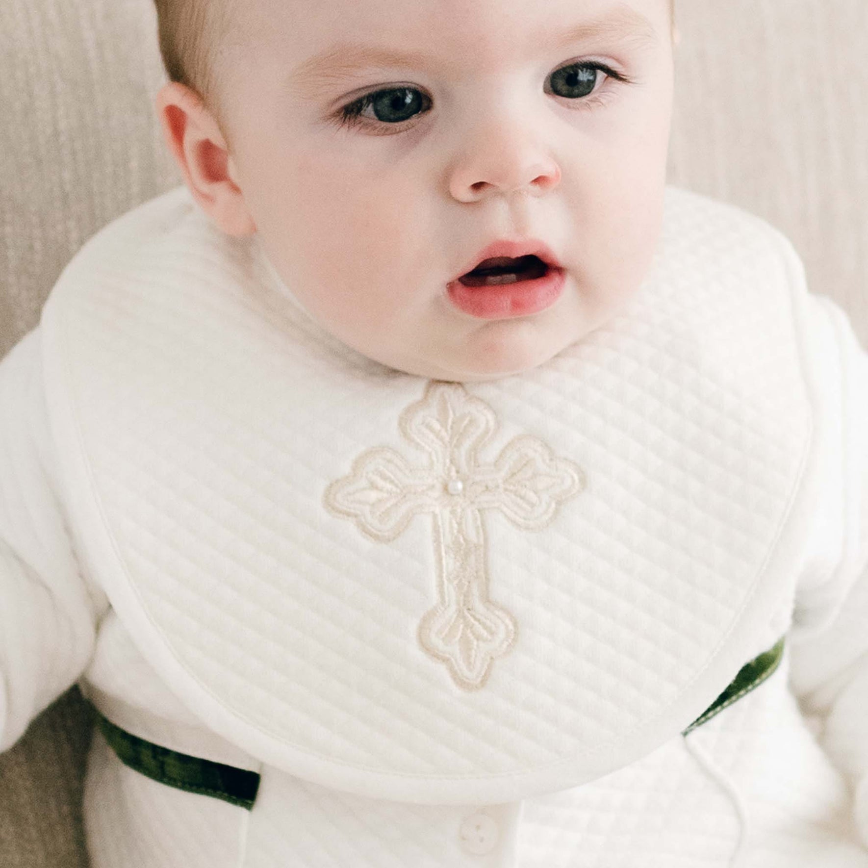 A baby wearing the Boys Cross Bib, adorned with an embroidered cross, sits on a beige surface and gazes slightly to the side. The baby is dressed in a white outfit with a touch of green peeking through.