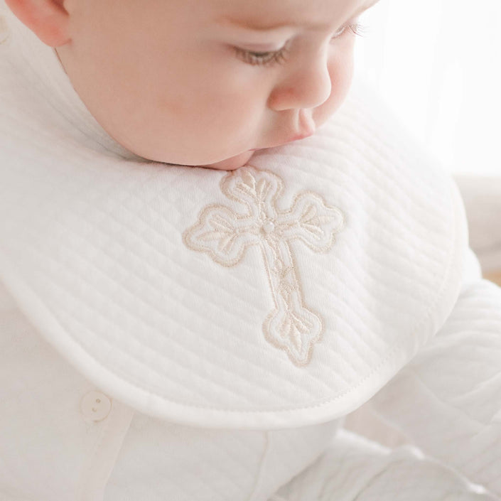 A baby is looking down while wearing a white quilted outfit with an embroidered cross on the bib, known as the Boys Cross Bib, handmade in the USA.