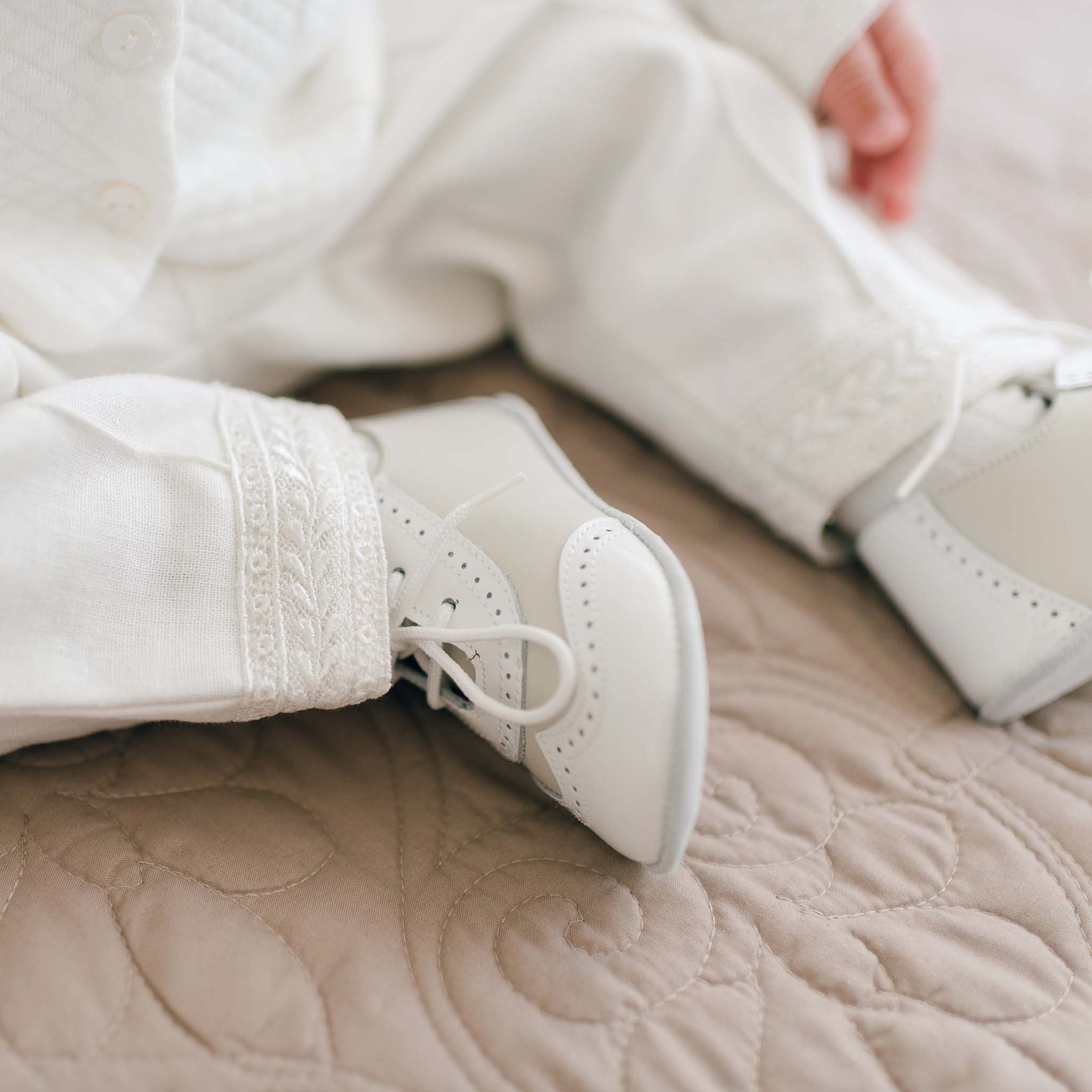 Close-up of a baby's legs and feet wearing the Ivory Two Tone Wingtip Shoes and the Oliver Ivory Suit Pants. The baby is sitting on a quilted beige blanket. Only part of the baby's outfit and a tiny portion of an arm are visible.