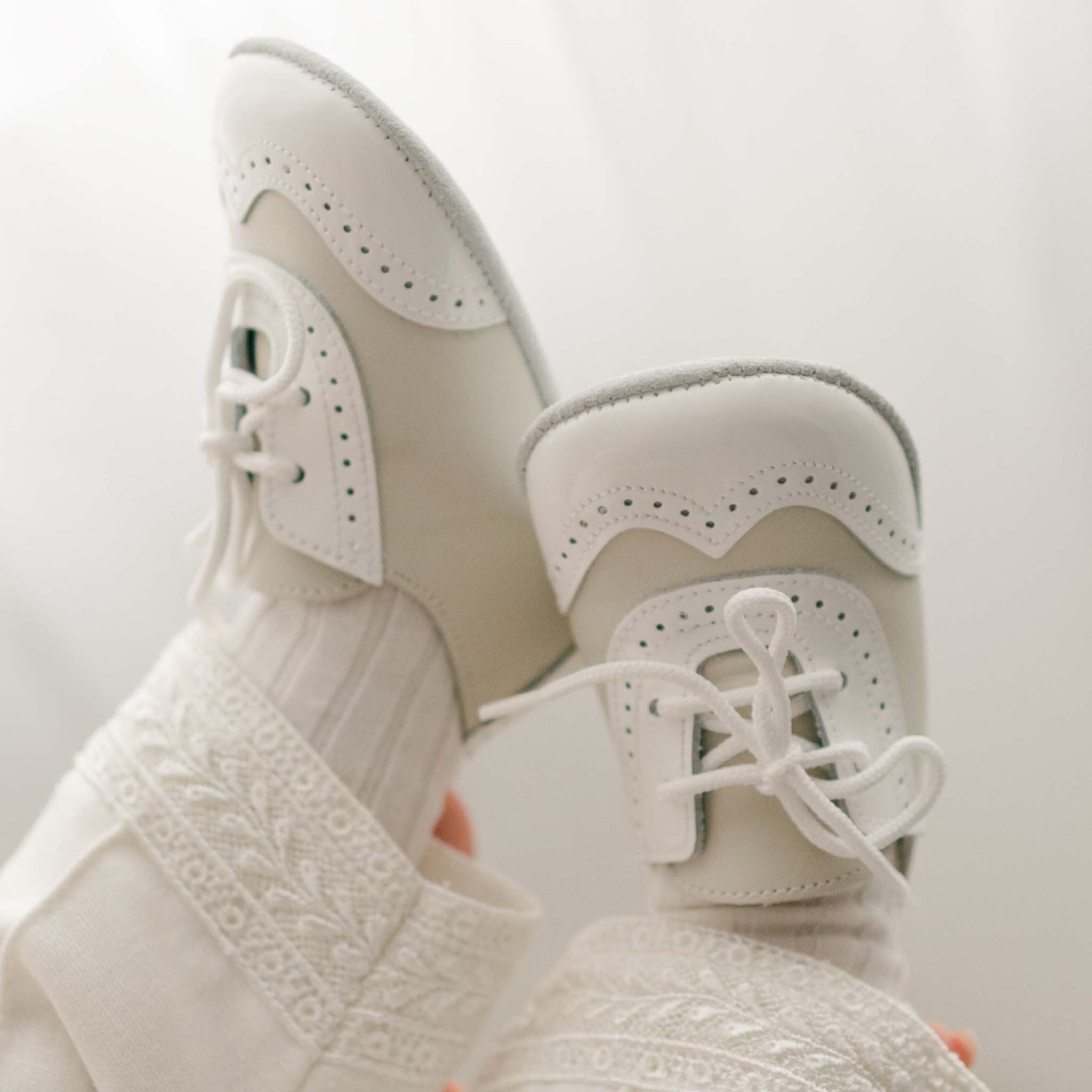 A close-up of a pair of Boys Ivory Two Tone Wingtip Shoes with laces and perforated detailing. The shoes are worn with textured white socks, creating an elegant look by Baby Beau & Belle. The background is softly lit with a neutral tone.