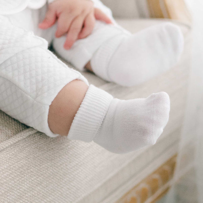 A baby's legs and feet are shown wearing quilted white pants and a pair of Boys Simple Socks, resting on a light-colored fabric surface. The baby's hand is visible near the leg, creating a setting that appears soft and comfortable.