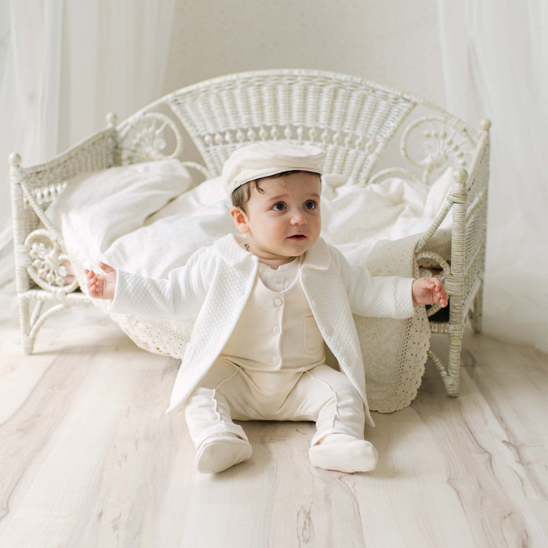 Baby boy in the Braden Coat Suit and matching Newsboy Cap sitting on a wooden floor with a white wicker chair in the background. Part of Christening Gowns' luxury baby clothes.