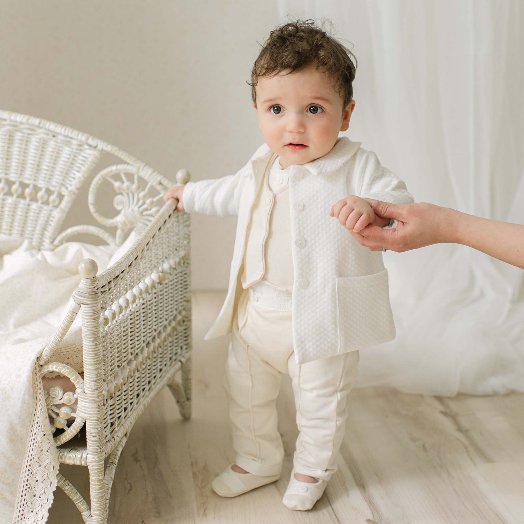 Baby boy in a Braden Coat Suit and matching Vest Suit standing next to a wicker chair in a bright room. Part of the Christening Gown Baby Boy Suit Collection.