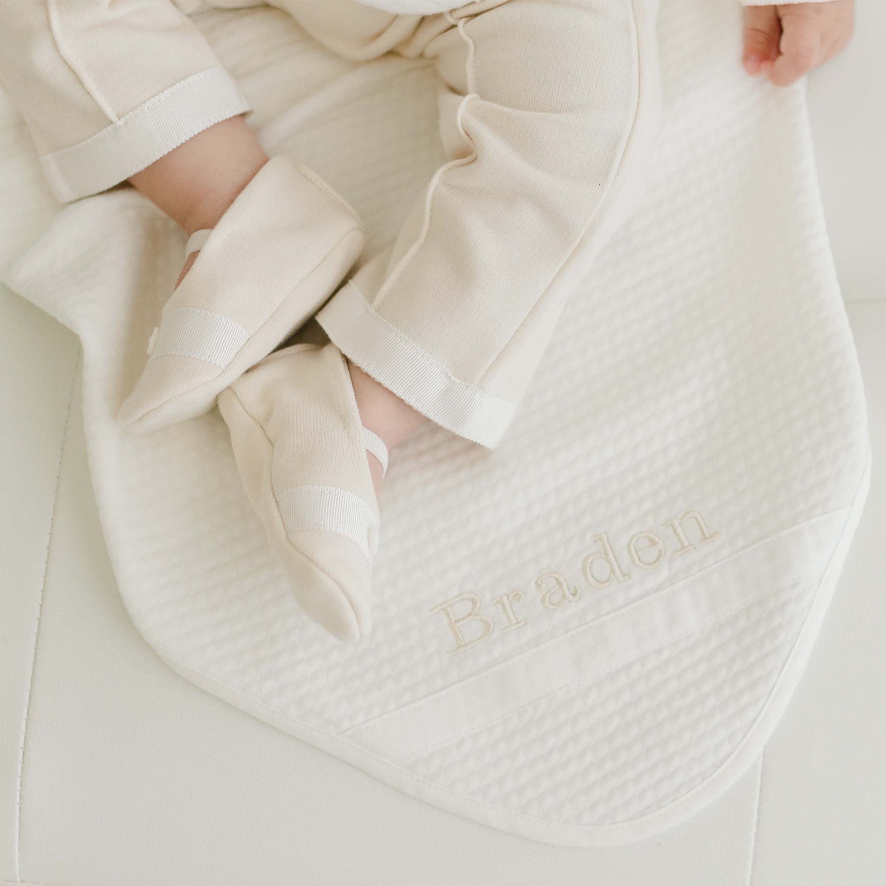 Baby laying on the Braden Personalized Blanket made from ivory textured cotton with an ivory grosgrain ribbon woven on the corner. The name "Braden" is embroidered on the corner of the blanket.