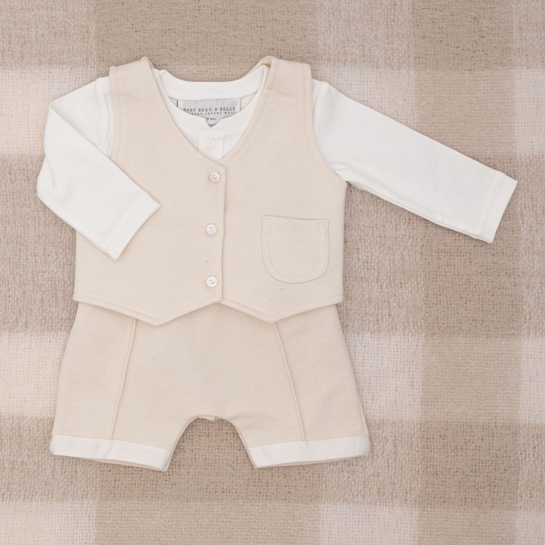 The Braden Vest Suit is a stylish and brand-new beige ensemble for babies, resembling a baby boy suit. It features a buttoned vest paired with shorts, all laid out on a checkered beige and cream blanket. Beneath the vest is a long-sleeved white shirt made from soft French terry, adding an elegant touch of formal wear for little ones.