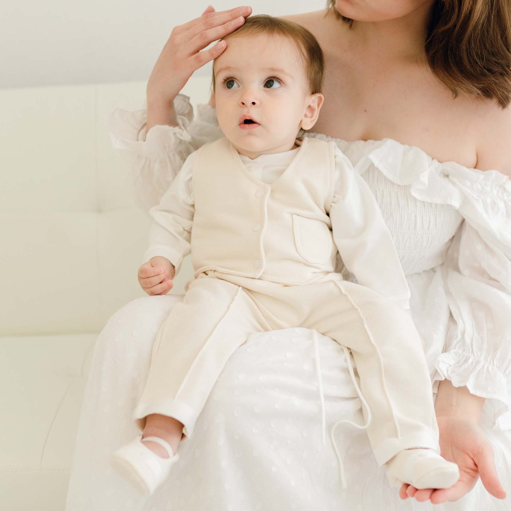 A woman in an off-shoulder white dress holds a baby boy dressed in the Braden Vest Suit on her lap on a white couch. The baby's cream-colored formal wear complements the cozy scene as she gently touches his head, her face softly hidden from view.