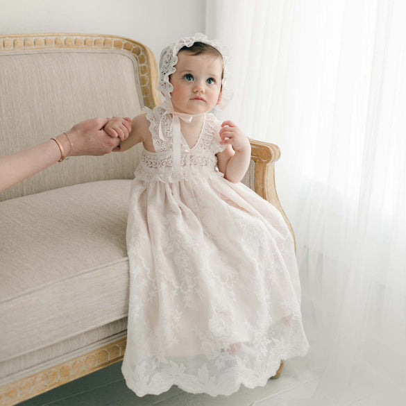 A toddler girl in a Charlotte Christening Gown & Bonnet sits on an elegant chair, looking curiously towards the camera with a slight frown. Her dark head is adorned with a matching lace bonnet.