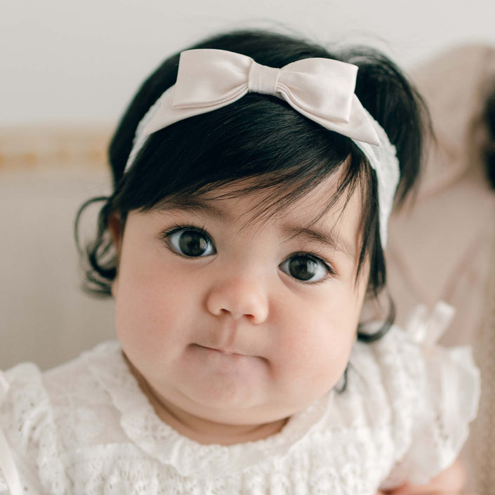 A baby girl wearing the Charlotte Lace Headband made with pink champagne silk and a floral lace. The baby looks directly at the camera with puckered lips.