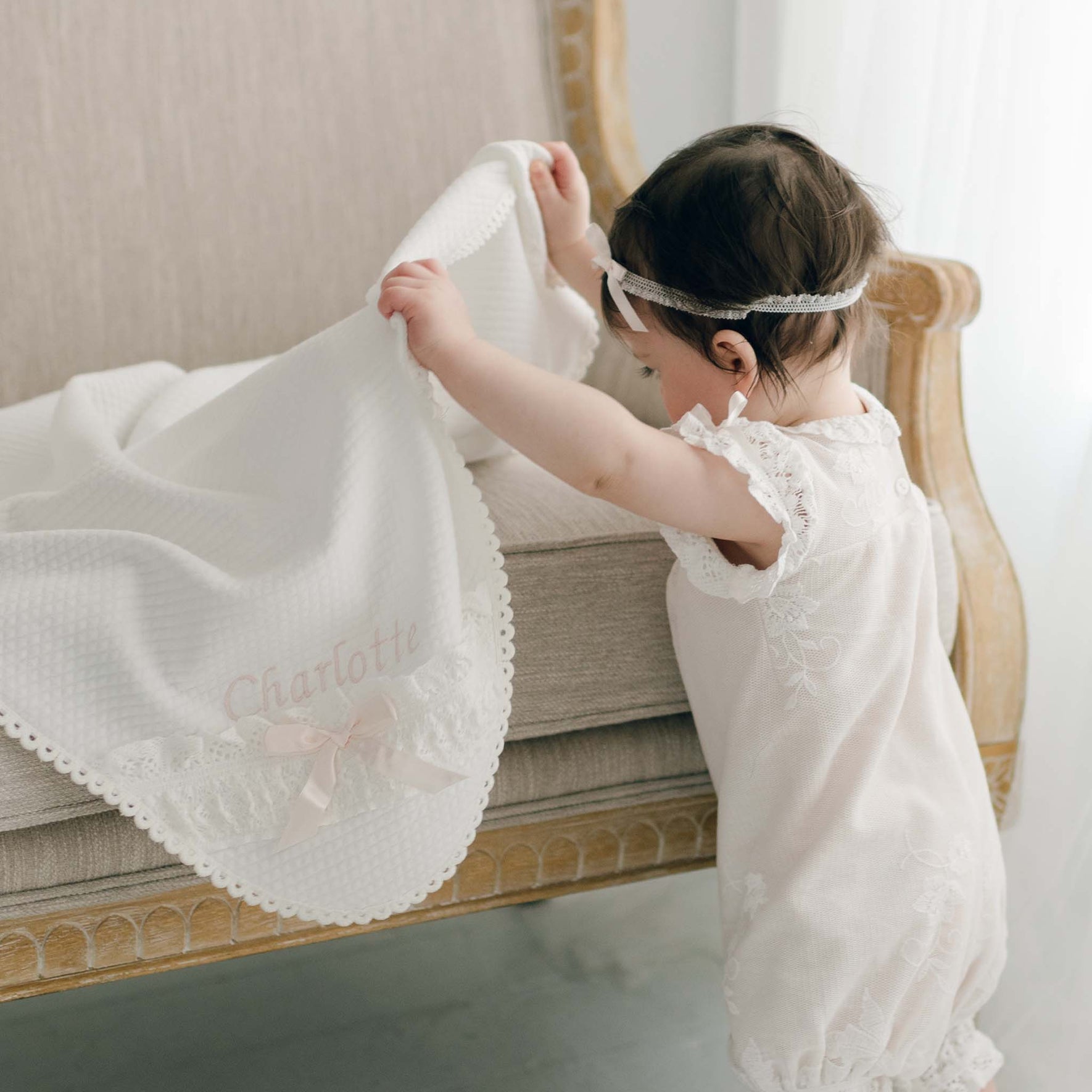 A toddler with dark hair, dressed in a lacy white outfit and headband, stands beside an ornate beige armchair. The child holds the Charlotte Personalized Blanket—featuring the name "Charlotte" embroidered on it and wrapped with a delicate pink silk ribbon.