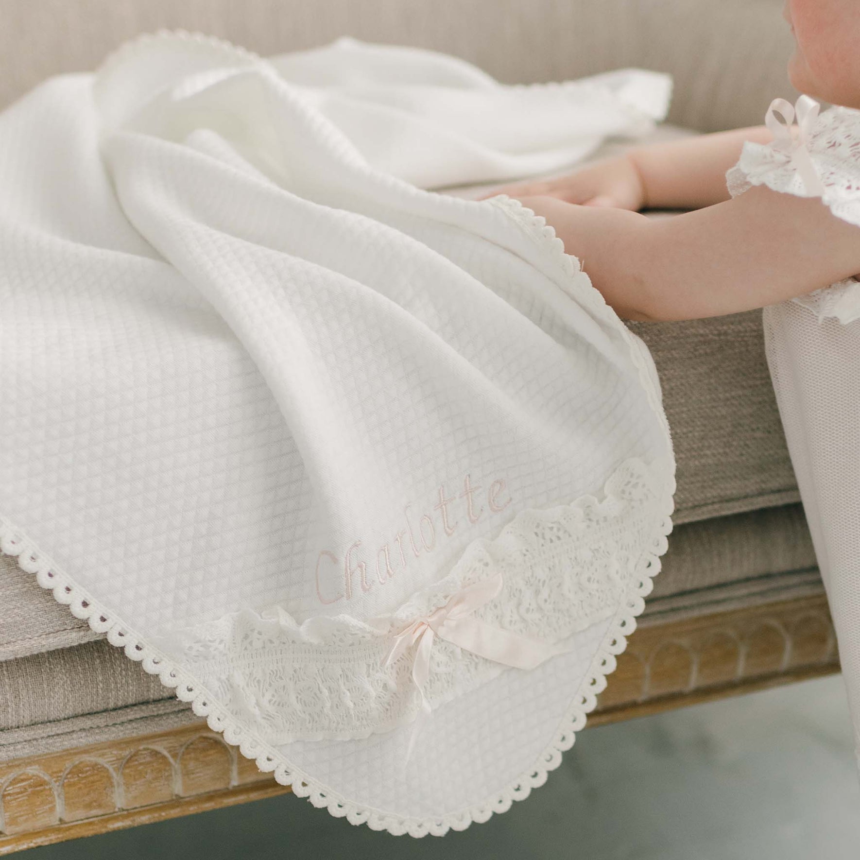 A baby is lying on a beige couch, partially covered by the Charlotte Personalized Blanket, an ivory quilted cotton blanket featuring lace edging. This keepsake is embroidered with the name "Charlotte" in soft pink thread and adorned with a delicate pink silk ribbon bow.
